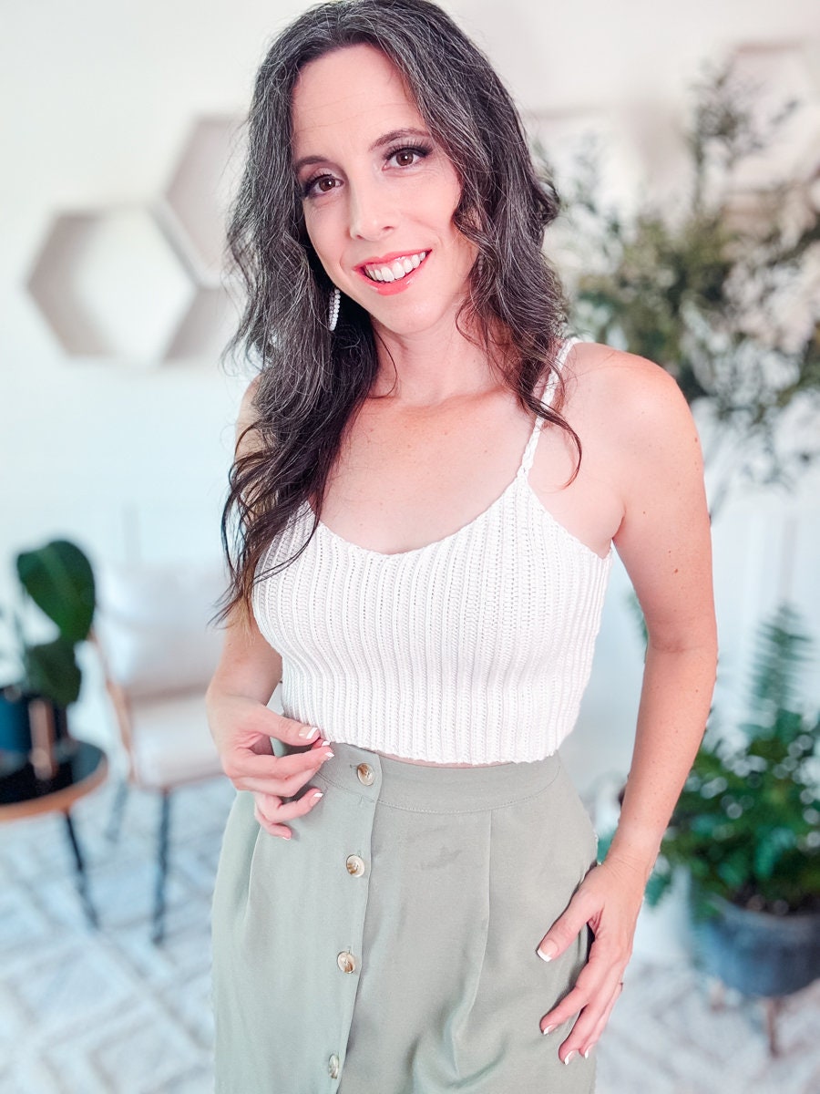 A woman in a white sleeveless crochet top and green skirt, accentuated by unique jewellery, poses indoors with a smile. She stands in front of a blurred background featuring plants and decorative wall elements.