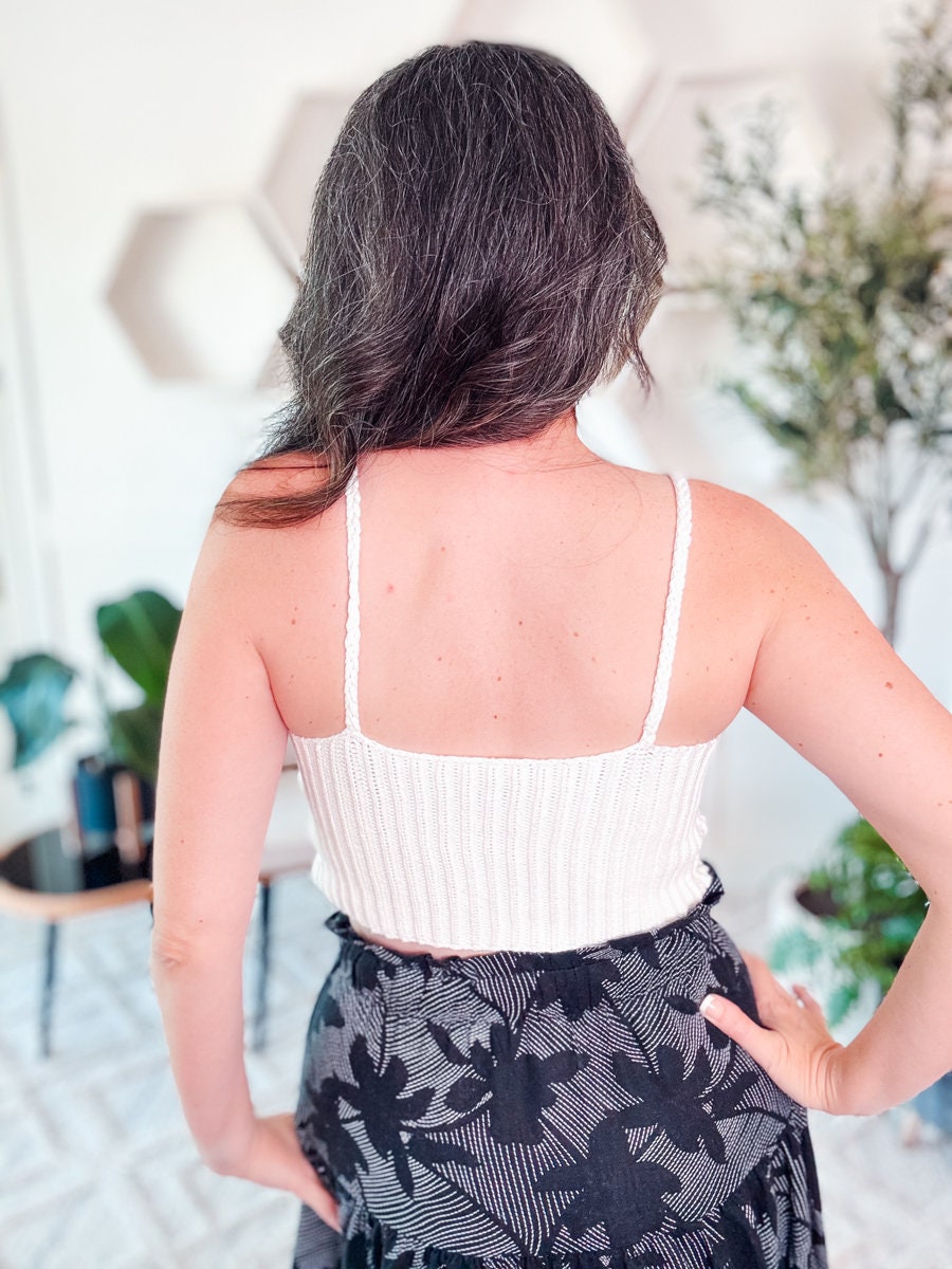 A person with long, dark hair with gray strands is seen from behind, wearing a white crochet tank top and a black skirt with a floral pattern. Adorning their neck is unique jewelry. The background includes plants and geometric wall decor.