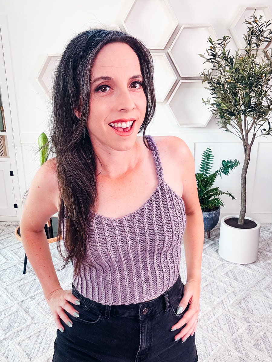 A woman with long dark hair wearing a purple crochet top and black pants stands indoors near a small green plant.