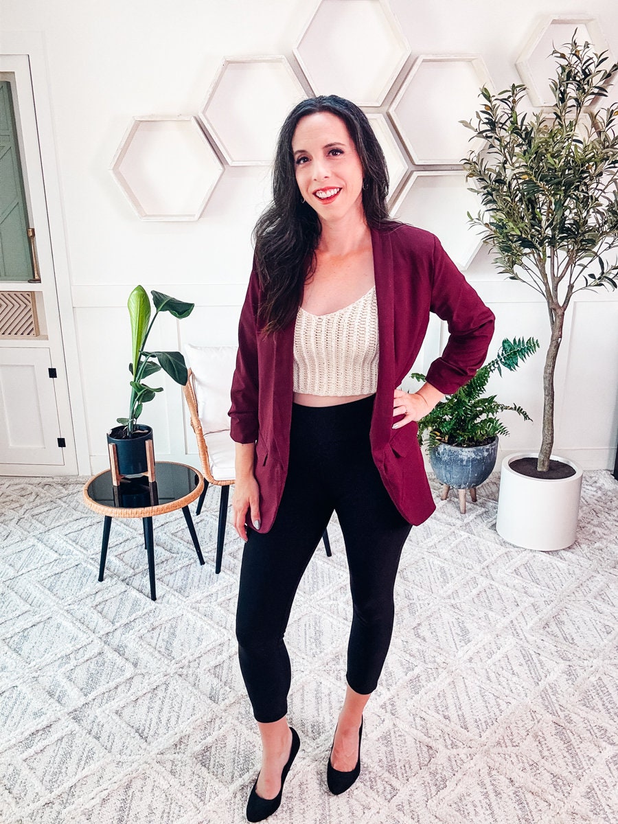 A woman stands indoors, smiling. She is wearing a maroon blazer, a crochet white top, black pants, and heels. Behind her are decorative hexagonal wall shelves, plants, a chair, and a door.