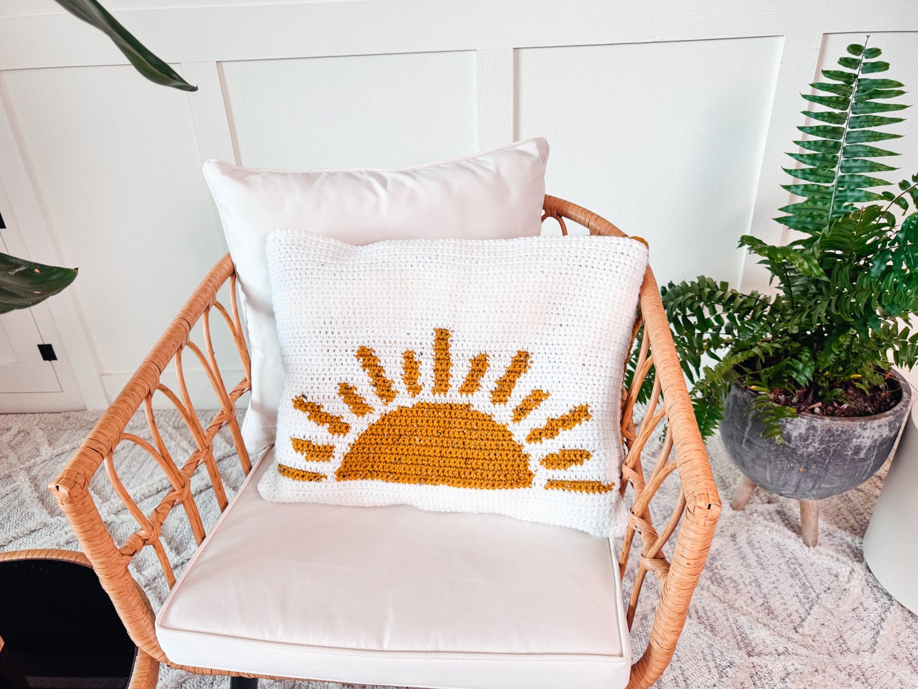 A rattan chair with white cushions, one featuring a crocheted pillow with a yellow sunburst design. Nearby is a green potted plant, creating an inviting nook perfect for showcasing unique jewellery.