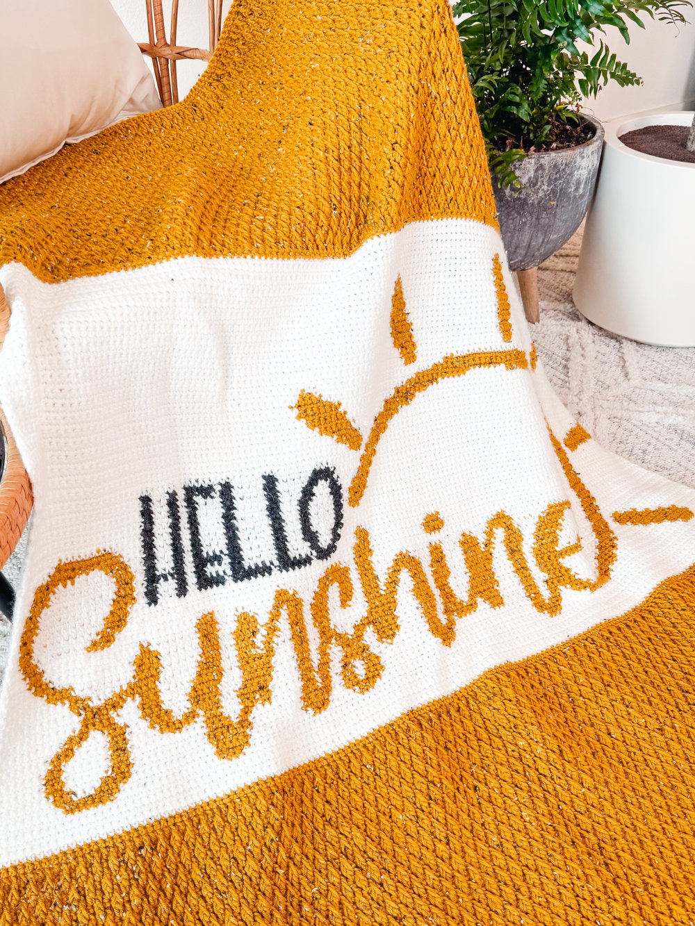 A knitted blanket with yellow and white stripes featuring the words "Hello Sunshine" and a sun motif. It is draped over furniture with green potted plants in the background, making it a perfect handmade gift.