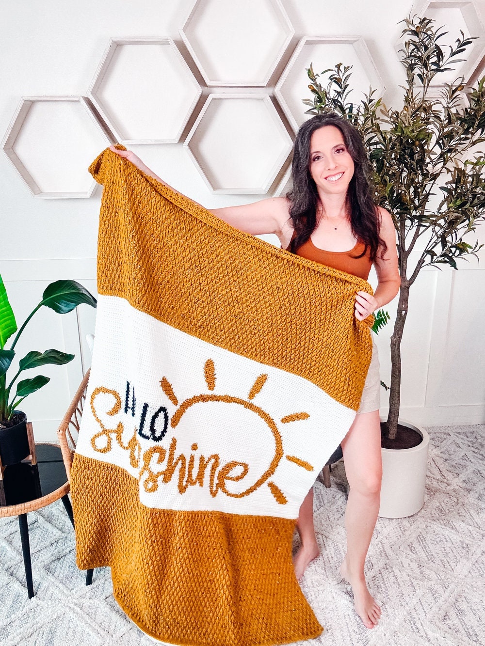 A woman stands indoors holding a yellow and white knitted blanket with the words "Hello Sunshine" and a sun illustration, perfect as a handmade gift. There are hexagon wall decorations and various plants in the background.