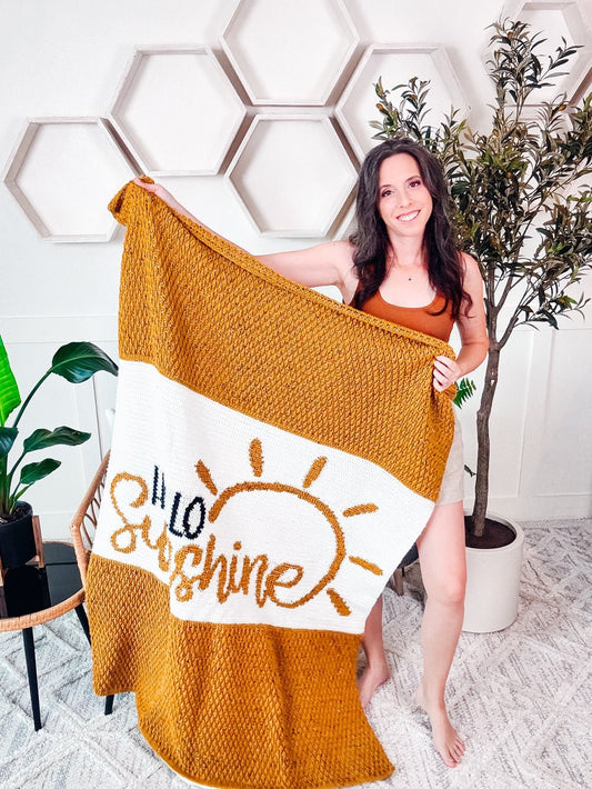 A woman stands indoors holding a yellow and white knitted blanket with the words "Hello Sunshine" and a sun illustration, perfect as a handmade gift. There are hexagon wall decorations and various plants in the background.