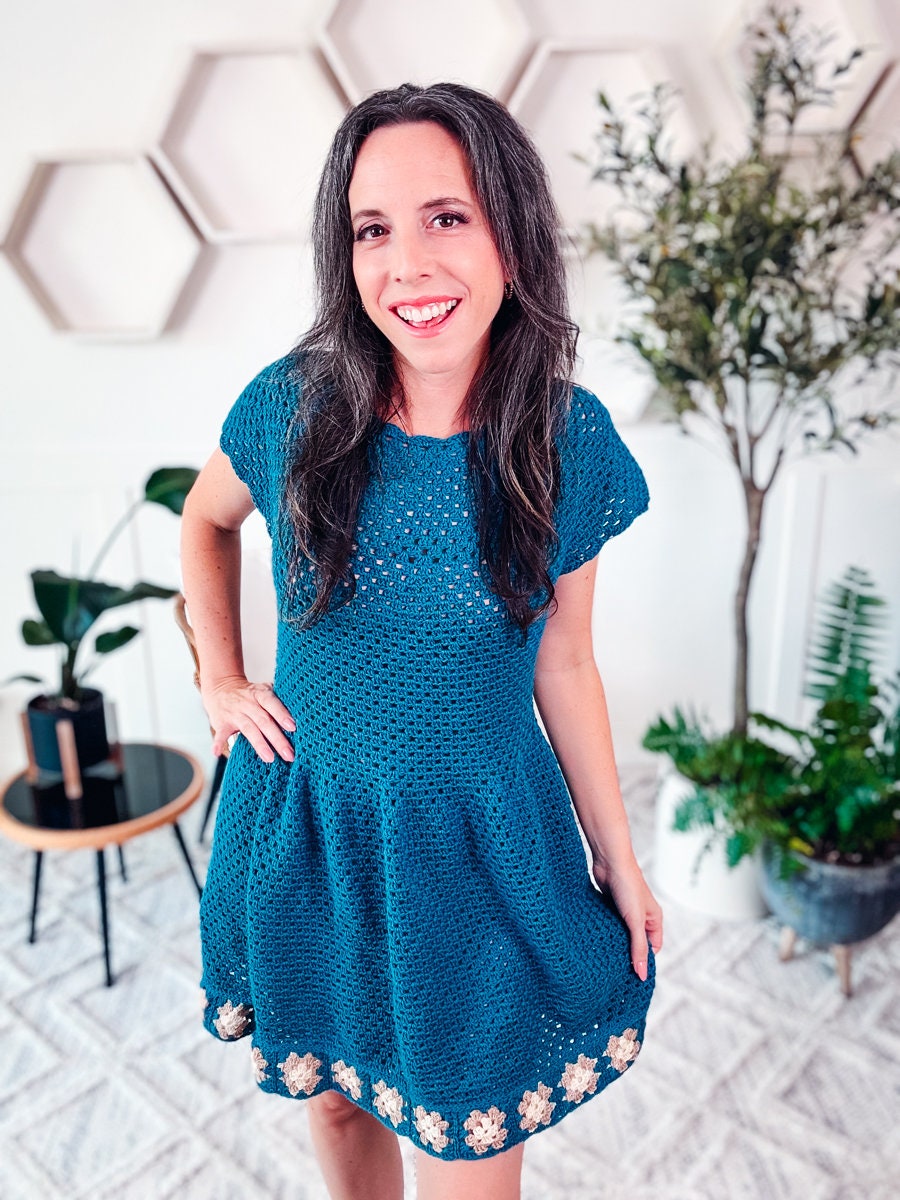 A person with long hair smiles while wearing a blue crocheted dress decorated with floral patterns at the hem, standing in a room with geometric wall decor, various potted plants, and displaying unique jewellery as a handmade gift.