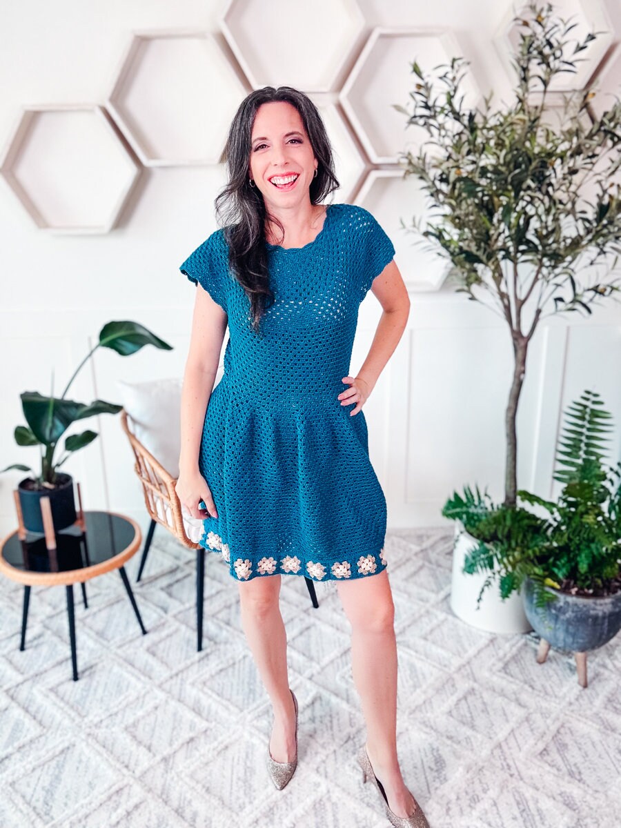 A person with long dark hair, wearing a blue dress and vintage clothes, smiling and standing in a room with hexagonal shelves, plants, and a geometric-patterned rug.