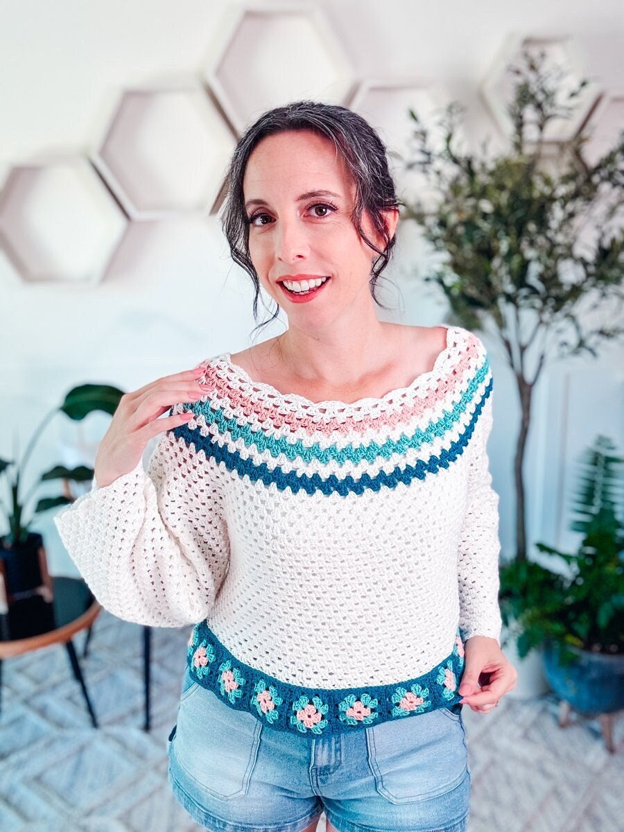 A woman stands indoors wearing a white crocheted top with colorful patterns on the collar and hem, pairing it perfectly with blue denim shorts. She smiles at the camera. The background features geometric wall decor and plants.