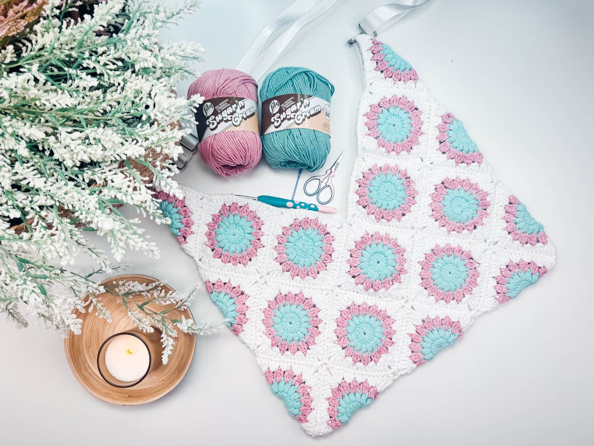 Crocheted blanket with pink and blue flower patterns alongside yarn, a crochet hook, and scissors. A lit candle and greenery are also present, making it a perfect handmade gift.