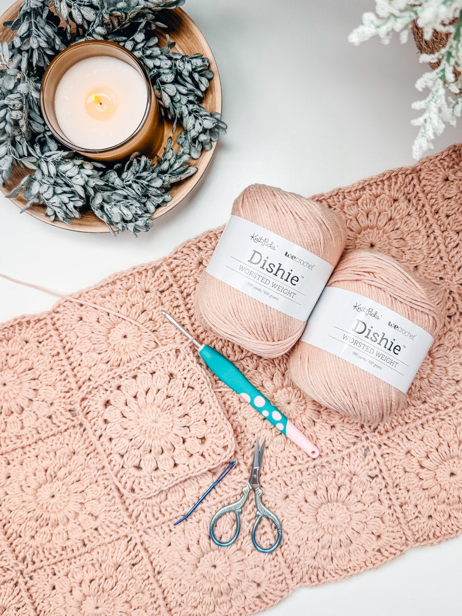 A crochet project with pink yarn, crochet hooks, scissors, and a candle in a decorative arrangement makes a perfect handmade gift.