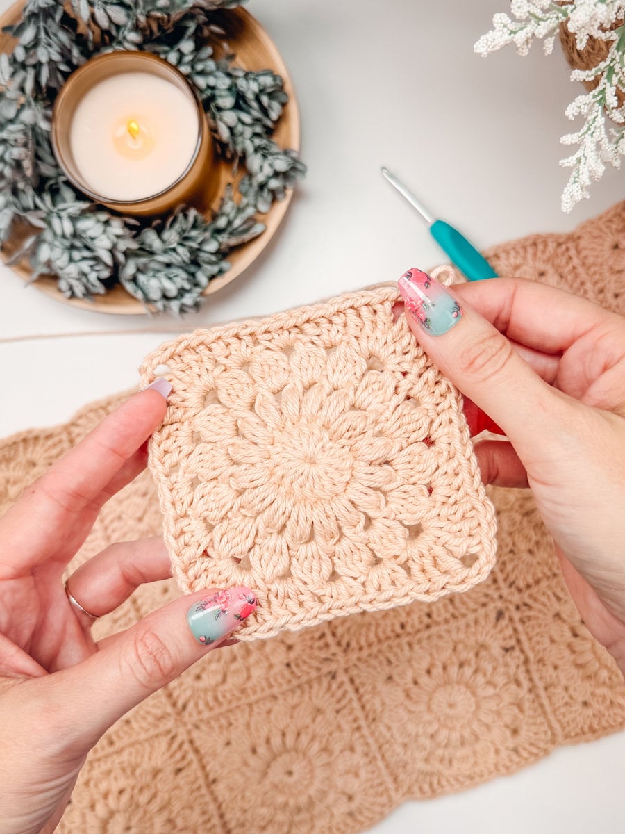 Hands holding a crocheted square with floral design, surrounded by crochet materials, candle, and greenery in the background—an ideal setting for creating a charming handmade gift.