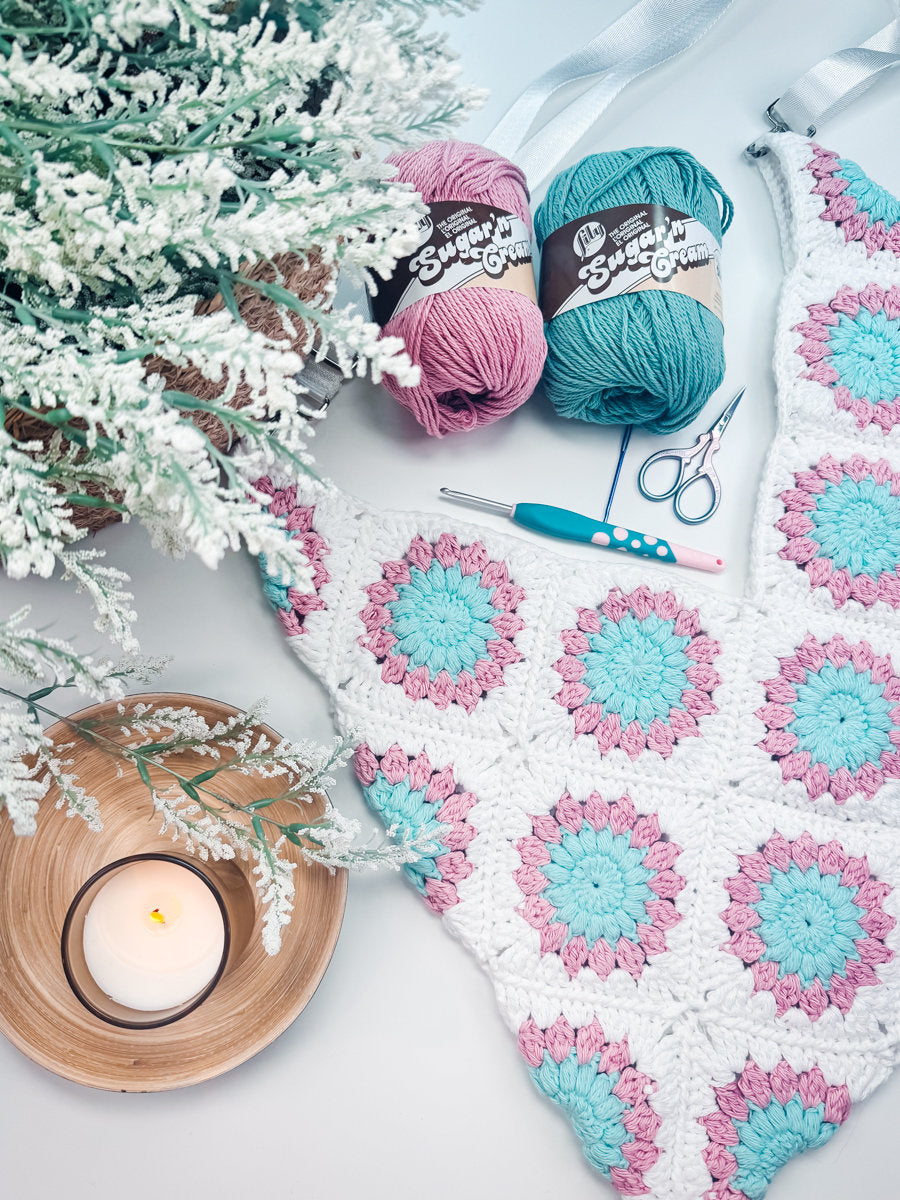 A crocheted piece with blue and pink floral motifs, two yarn skeins, a crochet hook, scissors, a small lit candle, and a frosted plant are arranged on a light surface—making it the perfect setting for crafting handmade gifts.