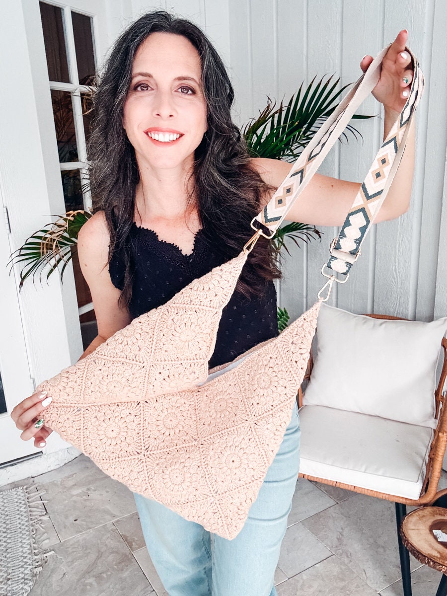 A woman with long dark hair holds a patterned beige bag with a decorative strap. She is standing indoors near a white door and a potted plant, her vintage clothes adding to the scene's charm.