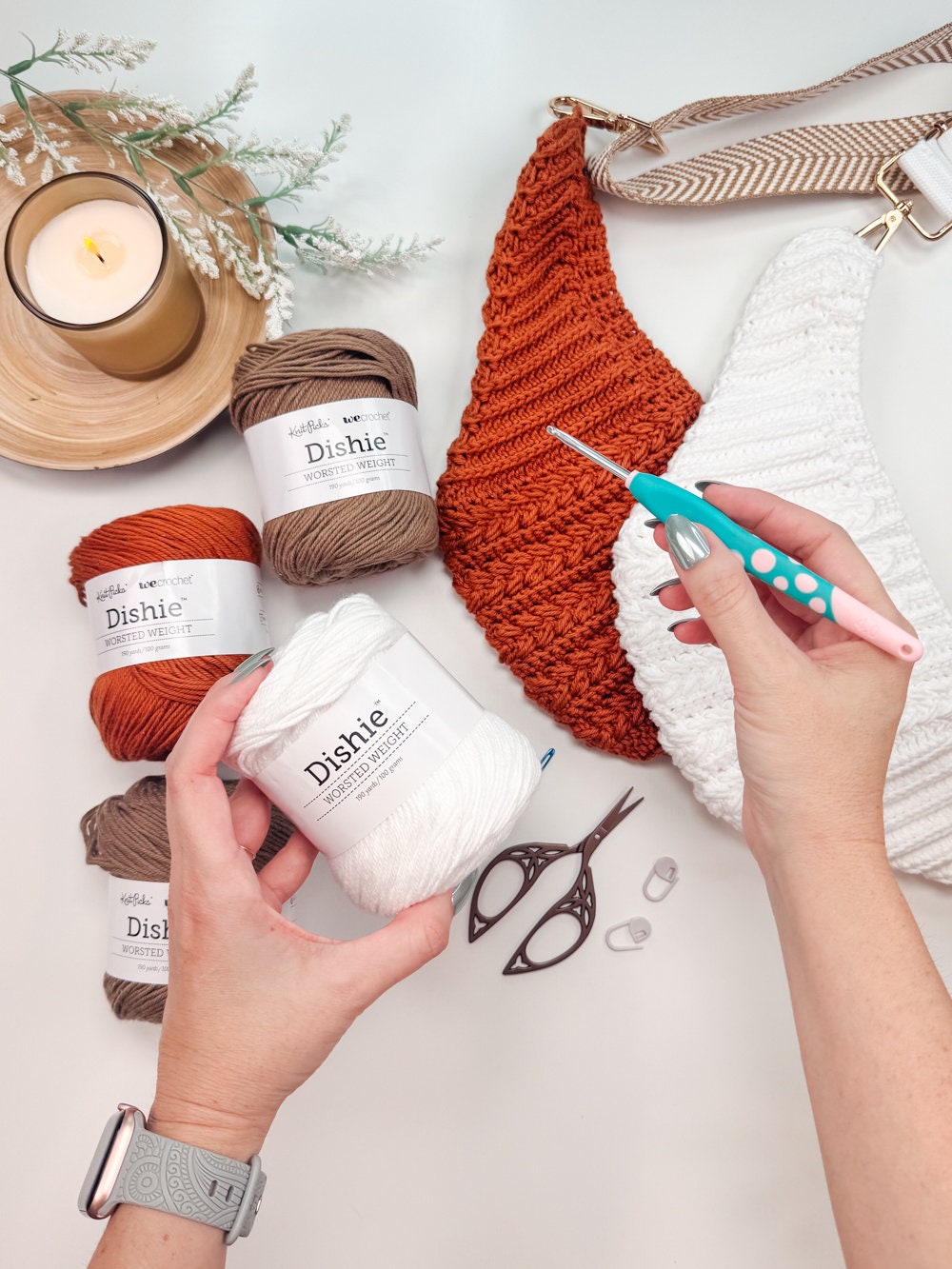 Hands holding a crochet hook near yarn, scissors, and partially completed crochet projects, with a lit candle and plant next to them—perfect setting for crafting a handmade gift.