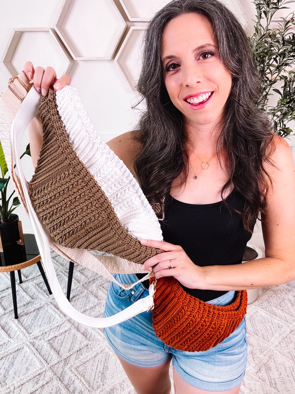 A woman with long, dark hair holds several woven fabric items in different colors. She is wearing a black top and denim shorts. The background features hexagonal wall decor, a plant, and displays of unique jewellery.
