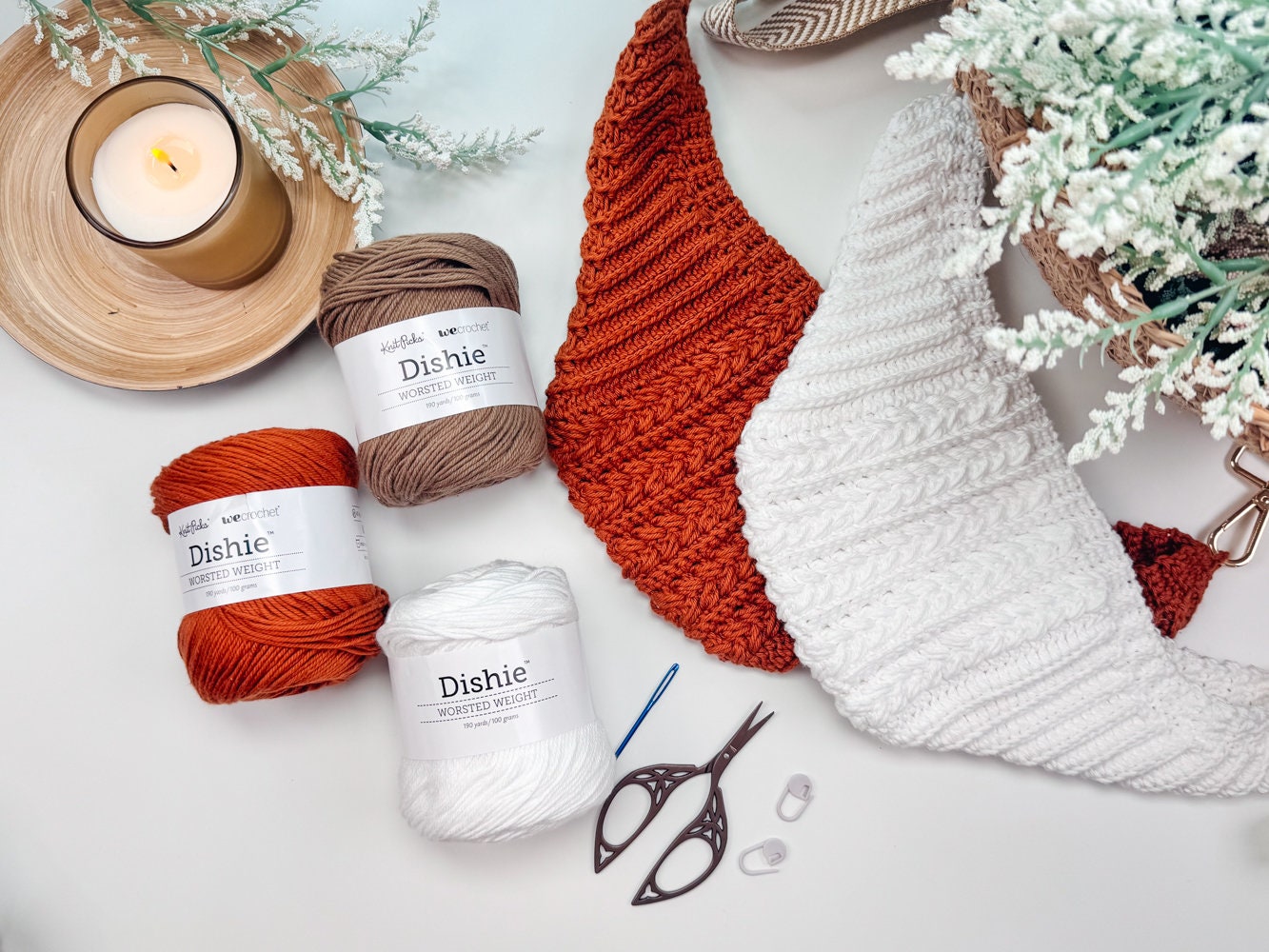 A knitting setup includes two scarves, a candle, three balls of yarn, a pair of scissors, and two needles arranged on a table, perfect for creating that handmade gift.