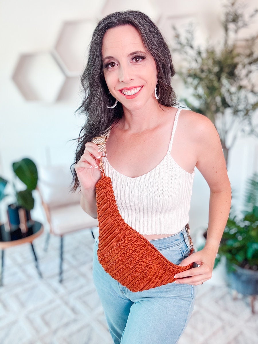 A woman smiles while holding a woven, orange handbag. She is wearing a white tank top and blue jeans, standing in a room with plants and geometric wall decor. Her vintage clothes and unique jewellery add to her stylish ensemble.