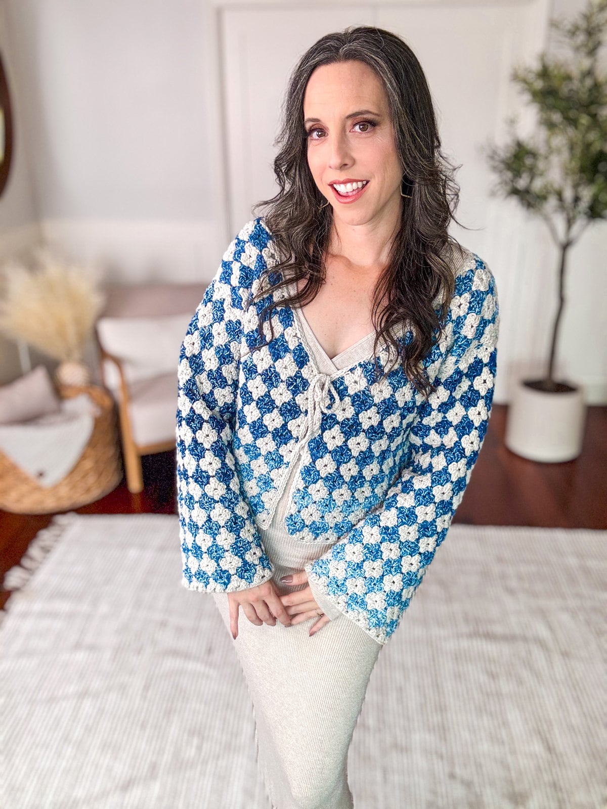 A woman with long dark hair, wearing a blue and white crocheted cardigan—a handmade gift—stands indoors on a light-colored rug with a plant in the background.
