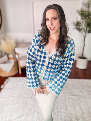 A woman with long dark hair, wearing a blue and white crocheted cardigan—a handmade gift—stands indoors on a light-colored rug with a plant in the background.