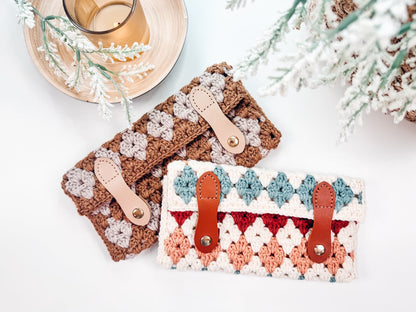 Two crocheted clutches with leather straps, one in brown and cream tones, the other in a mix of blue, red, and peach, are displayed on a table with a candle and greenery decoration. These handmade gifts pair perfectly with an effortlessly chic look.