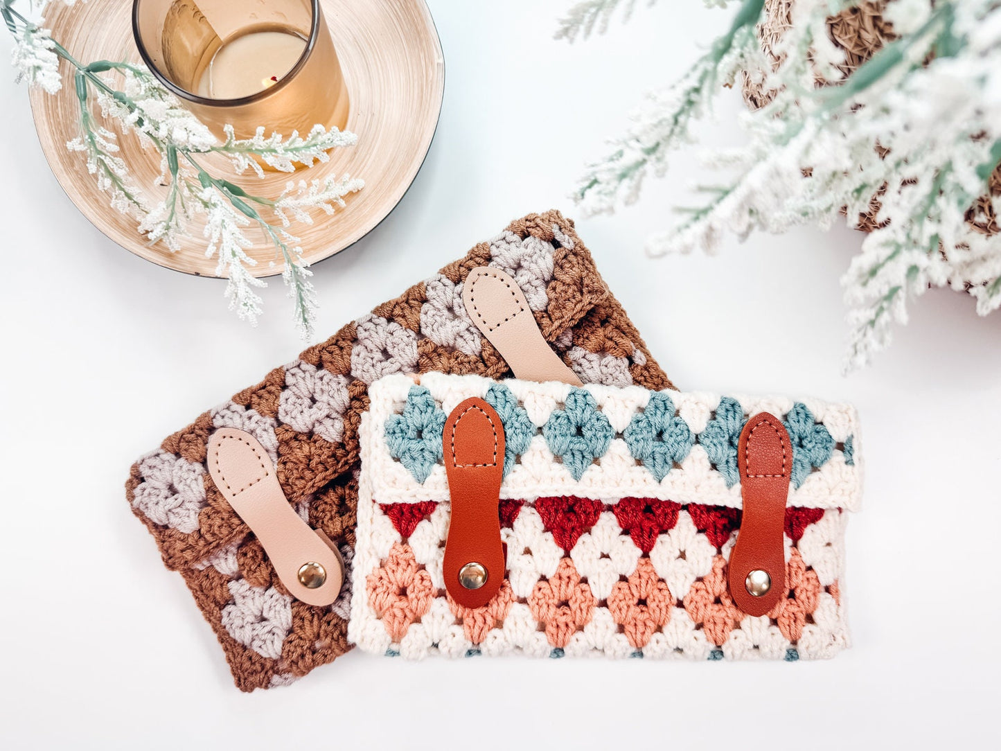 Two crocheted wallets with leather straps are placed on a white surface near a glass candle holder and frosted evergreen branches, offering a perfect handmade gift for anyone who appreciates unique jewelry.