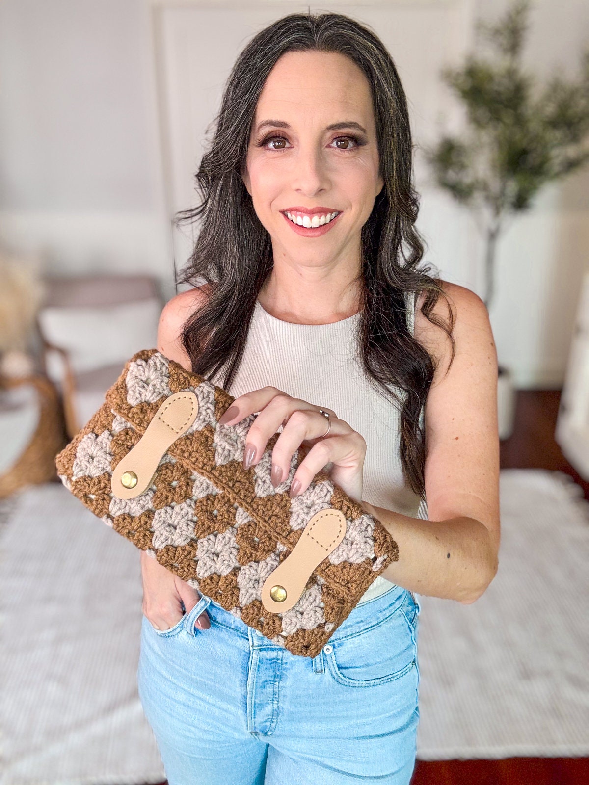 A woman standing indoors holds a crocheted handbag with a multicolored pattern, wearing a sleeveless top and jeans, smiling at the camera, accessorized with unique jewellery.