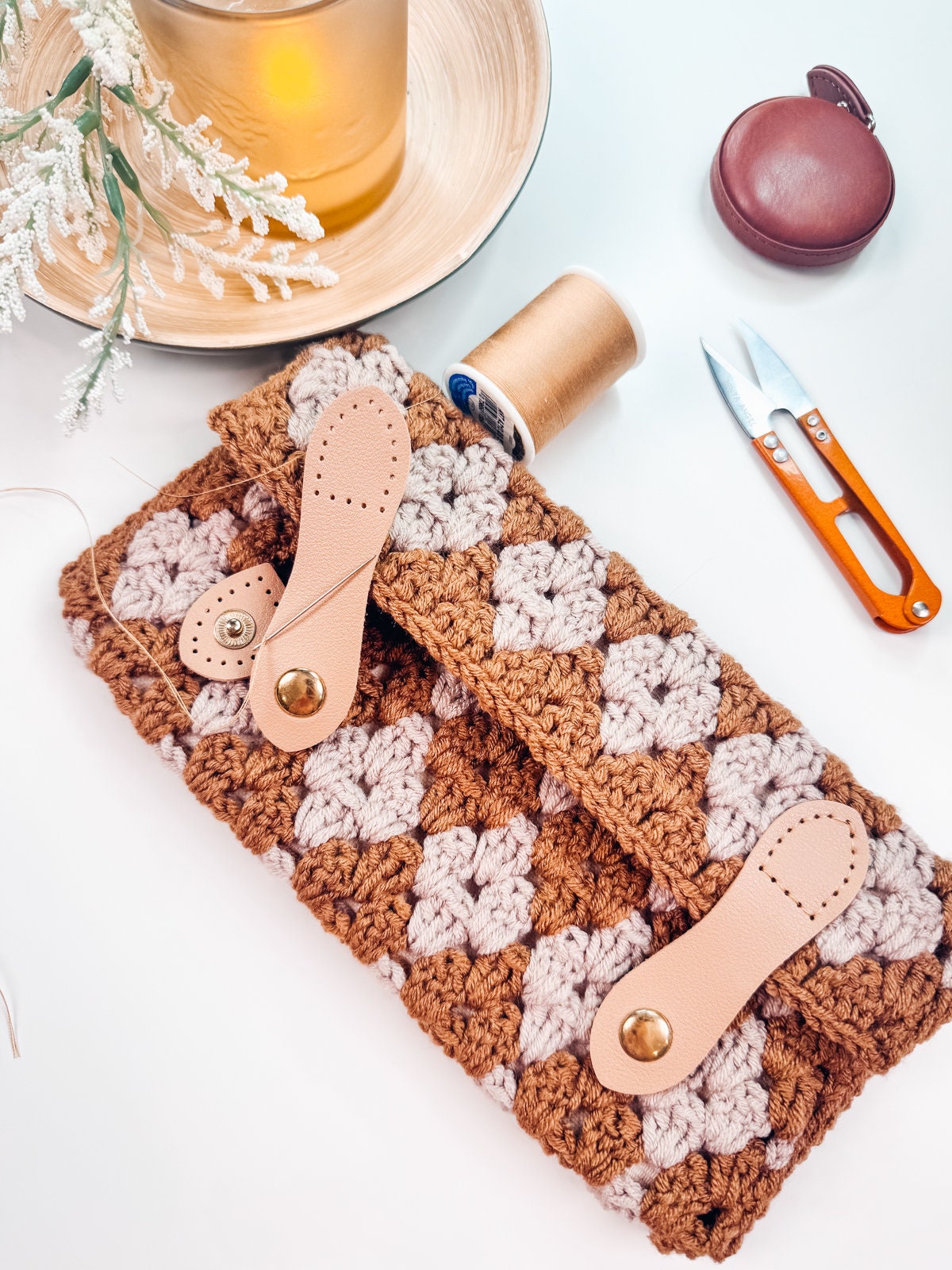 Close-up of a hand-crocheted clutch bag in brown and beige with leather straps on a table, surrounded by a candle, thread spool, compact case, small scissors, and unique jewellery. This handmade gift adds charm to any setting.