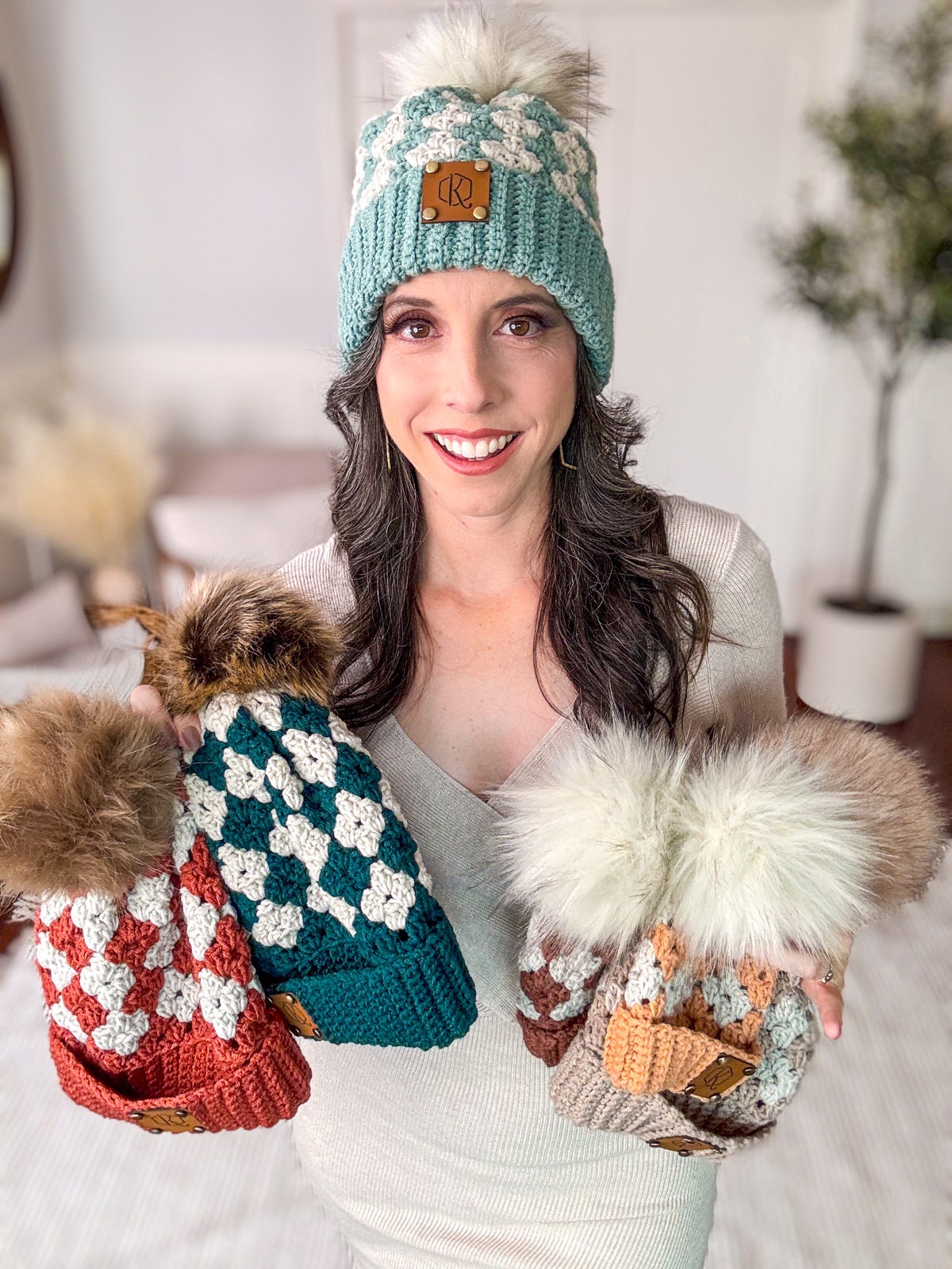 Woman smiling, wearing a light blue knit beanie with a pom-pom, holding three similarly styled, handmade gift beanies in different colors, in a well-lit indoor setting.