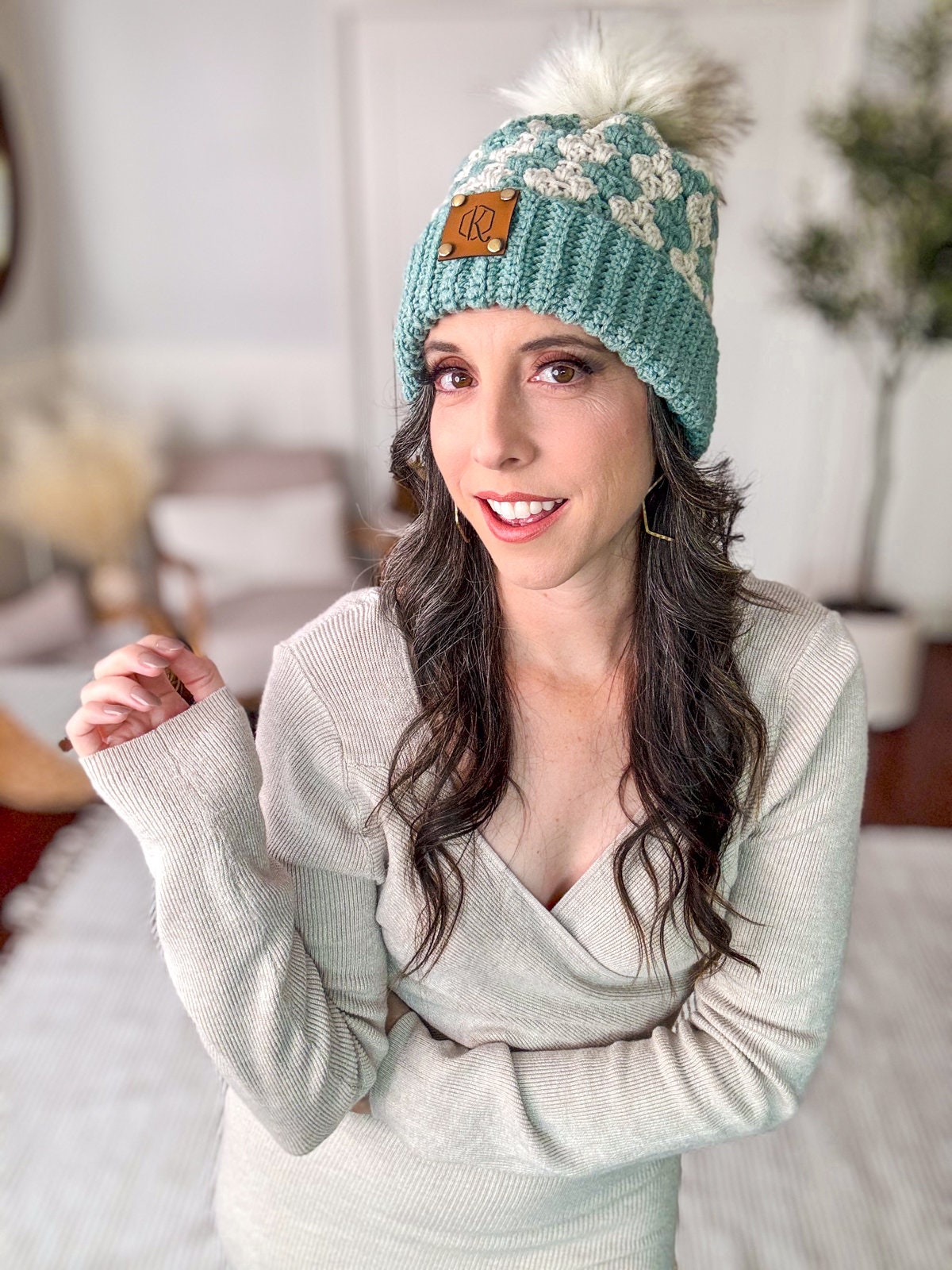Person wearing a green knit hat with a pom-pom and a light-colored sweater, standing in a room with vintage clothes, unique jewelry, and a couch with a plant in the background.