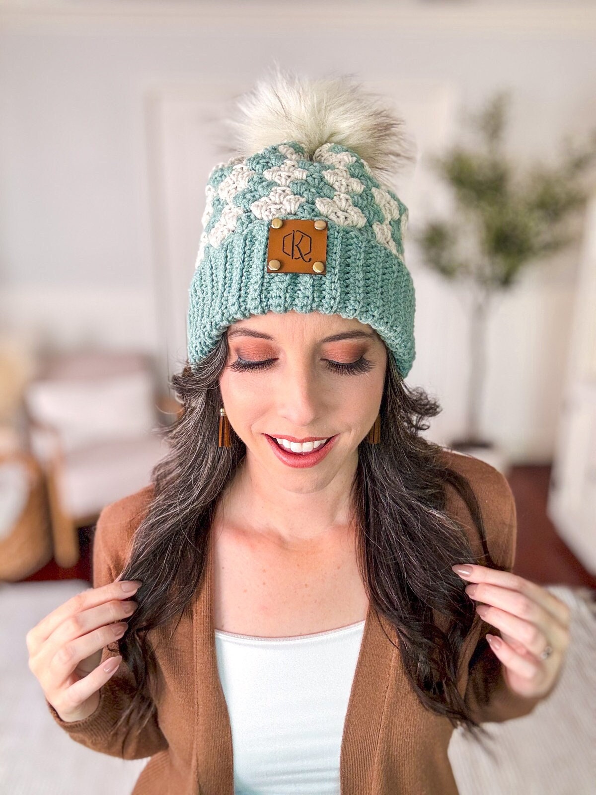 A person with long hair wearing a light blue knit beanie with a pom-pom and a brown cardigan, standing indoors, accessorized with unique jewellery.