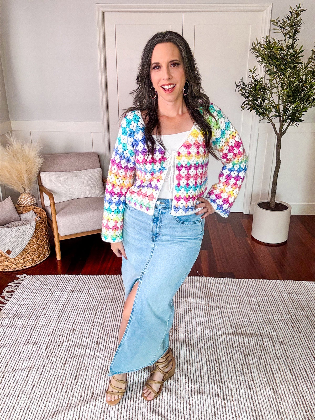 A woman stands indoors, smiling, wearing a multicolored crochet sweater, white top, and a long denim skirt with a front slit. She has wavy hair and is in a room with a chair, basket, and potted plant.