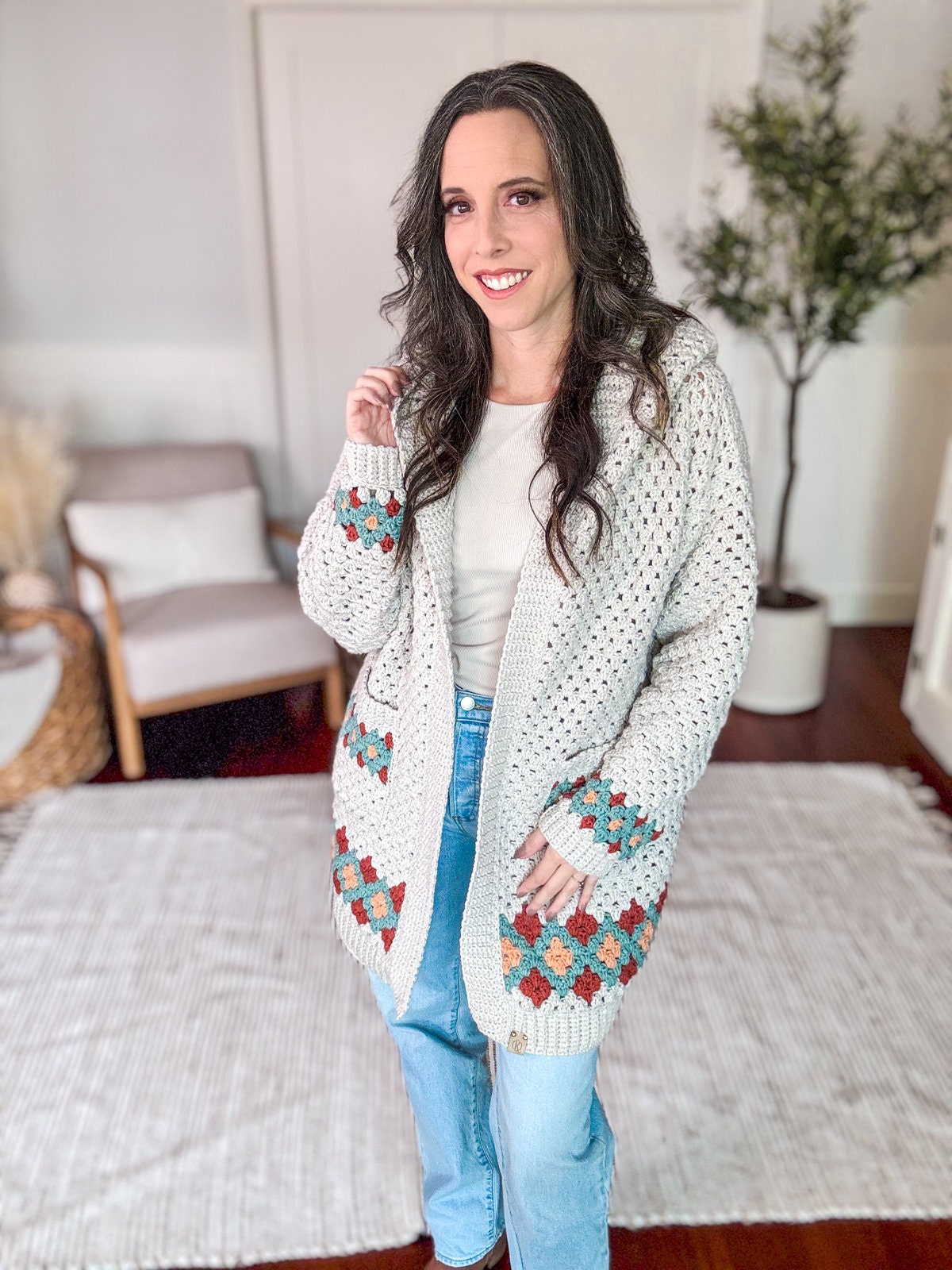 A woman with long, dark hair is standing indoors, wearing a white crocheted cardigan with colorful patterns, a white top, and light blue jeans. She is smiling and has her hands in her cardigan pockets, exuding charm.