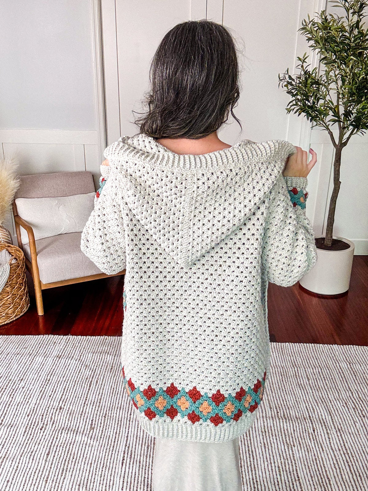 Person standing indoors with their back turned, wearing a white knit hoodie featuring a colorful pattern near the bottom. A small indoor plant and a woven basket chair are in the background.