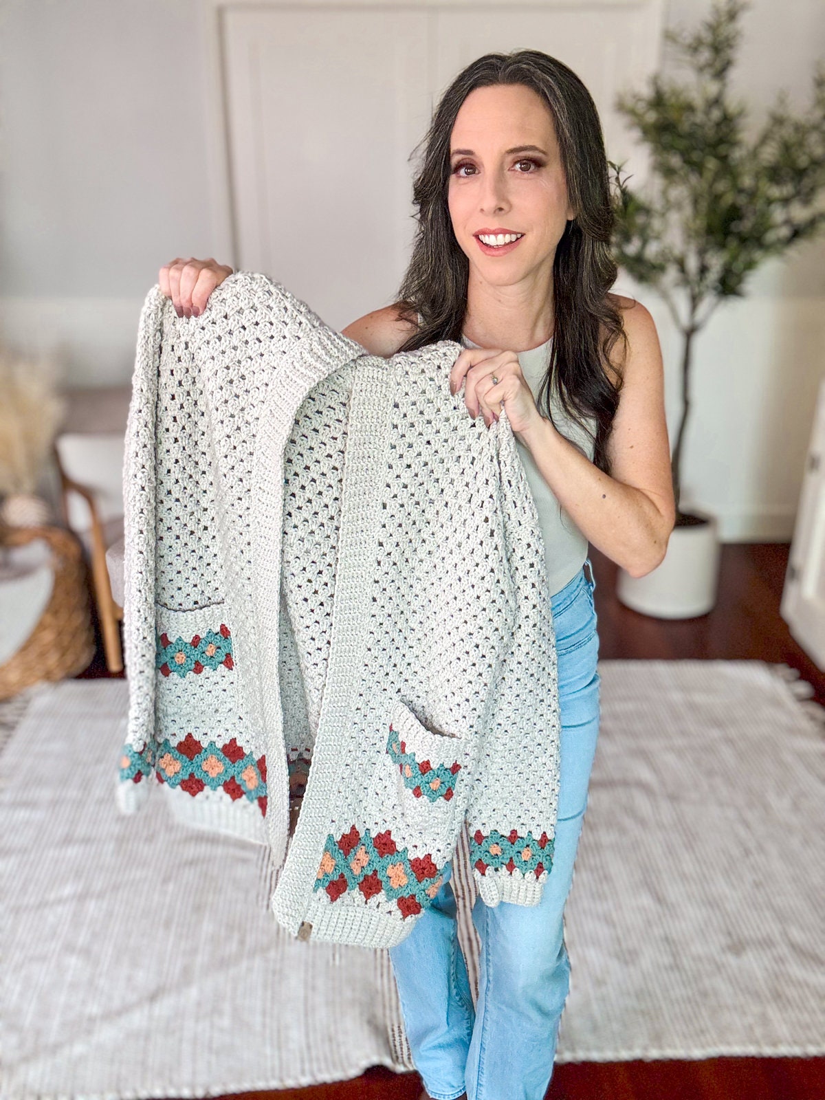 A woman holding a crocheted cardigan with colorful pocket details, standing indoors on a white rug with a green plant in the background, making for the perfect handmade gift.