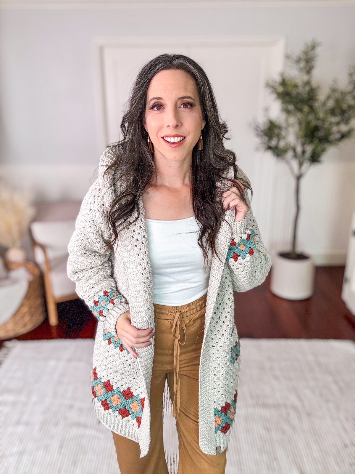 A woman wearing a knitted cardigan with colorful patterns and unique jewellery stands in a light-colored room with a small plant in the background.