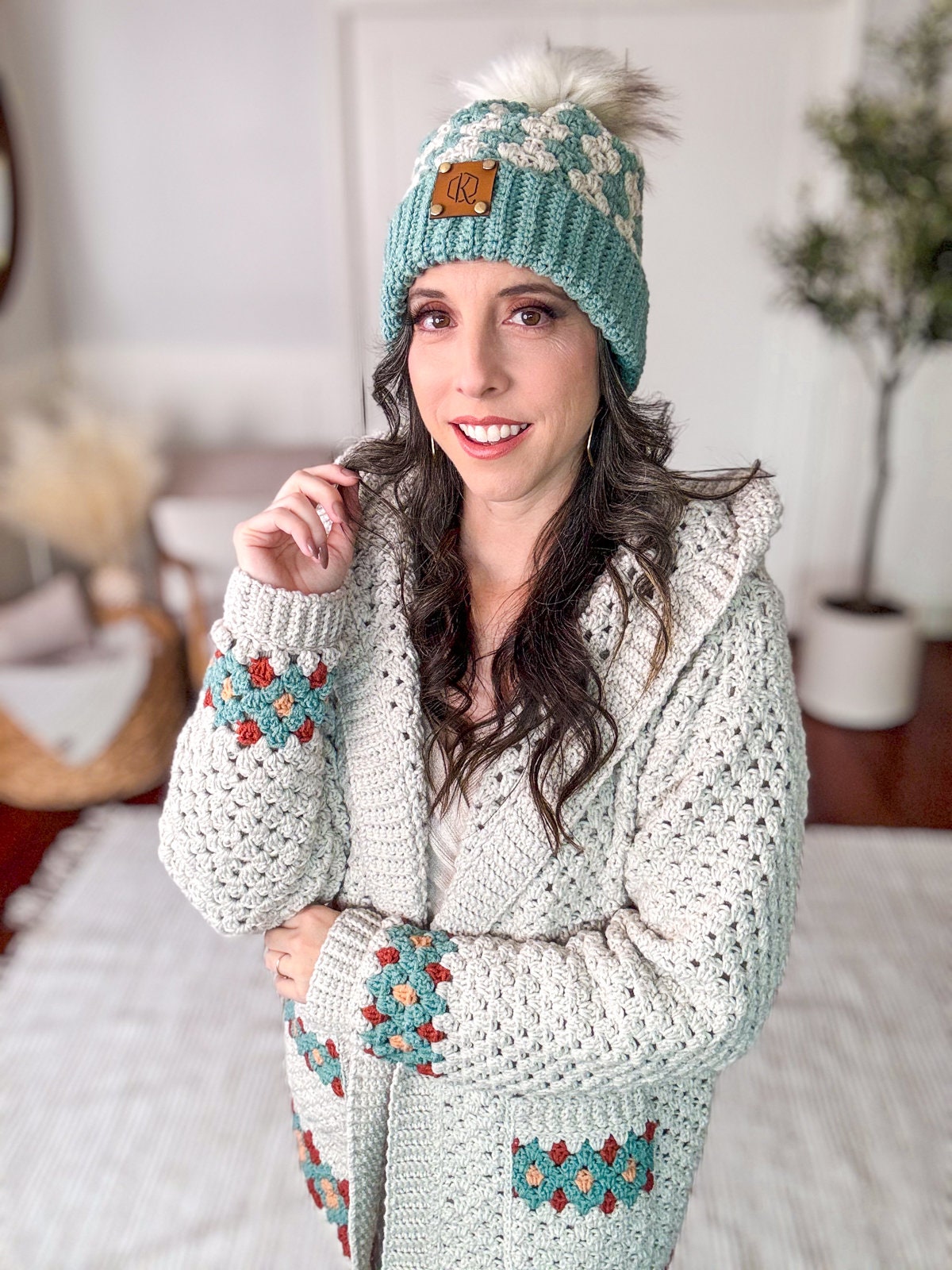 A person wearing a crocheted cardigan with colorful patterns and a beanie with a pom-pom, complemented by unique jewellery, is posing indoors.