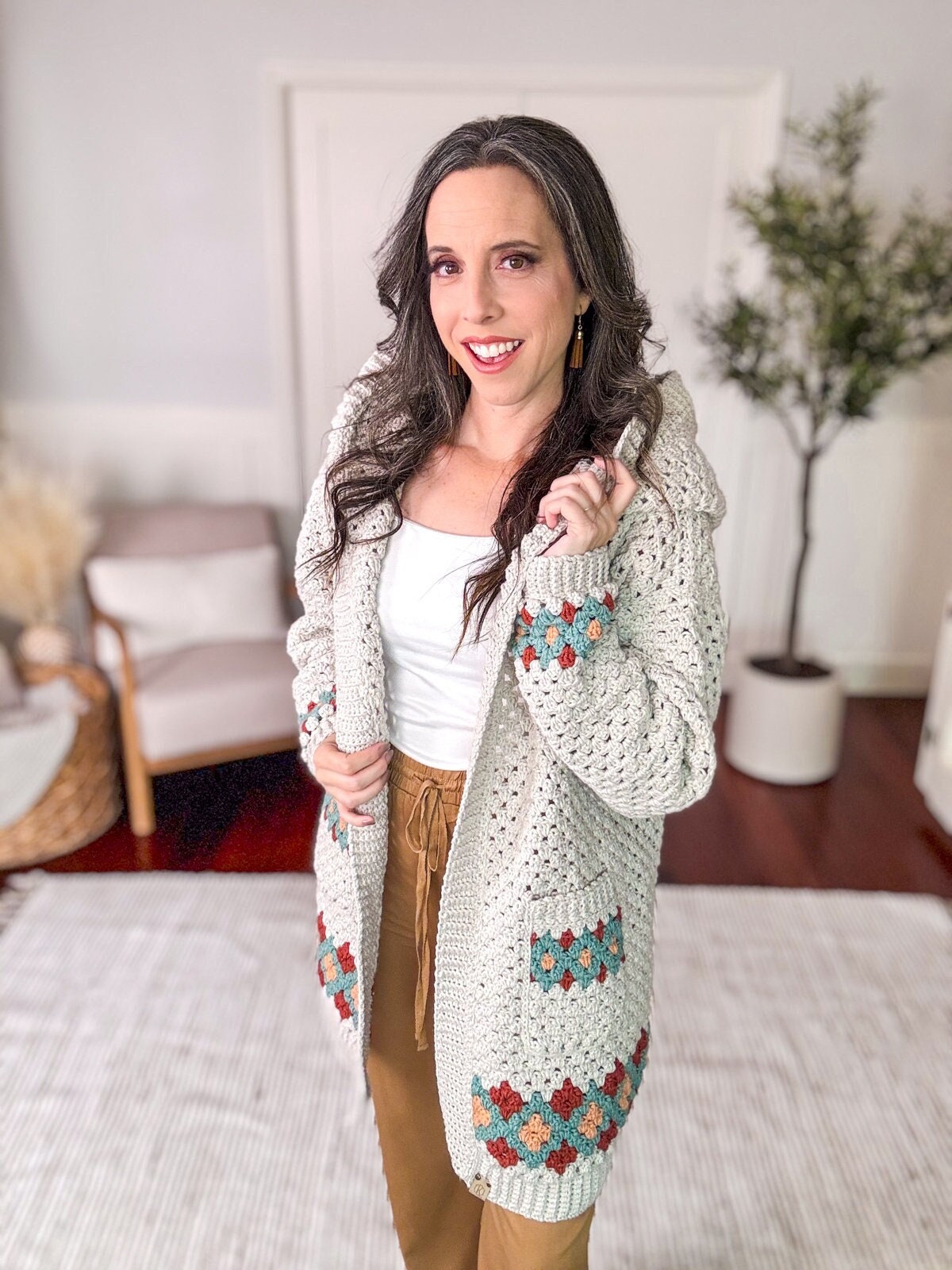 A woman in a white top and brown pants is smiling and wearing a knitted cardigan with patterned pockets, accessorized with unique jewelry. She stands in a well-decorated room with a plant and chair in the background.