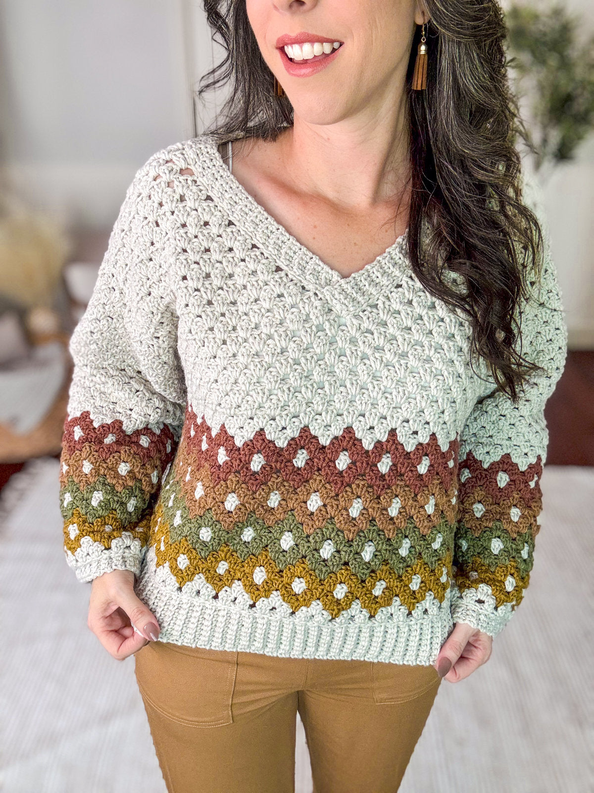 A person is wearing a crocheted sweater with a V-neck, featuring a white top section and a patterned design in brown, red, green, and yellow colors. Complementing their vintage clothes look, they are also wearing brown pants.