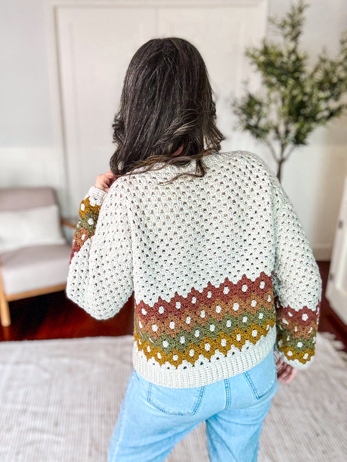 A person with long hair is wearing a crochet sweater with a multicolored pattern and blue jeans, adorned with unique jewellery, standing in a room with a chair and a plant in the background.
