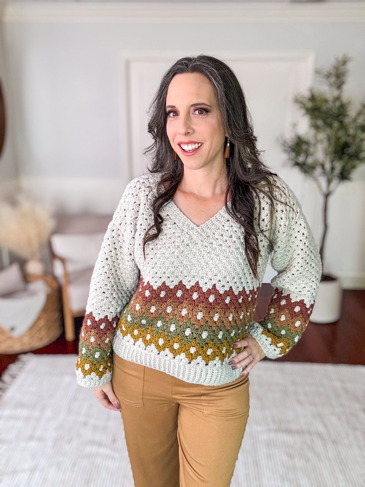 A person with long dark hair smiles while standing indoors. They are wearing a knitted sweater with a geometric pattern, brown pants, and unique jewellery that complements the vintage clothes. A plant and handmade gift can be seen in the background.