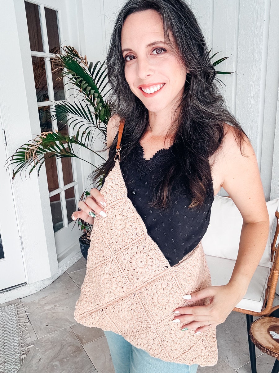 A smiling woman with long dark hair is wearing a black sleeveless top and a beige crocheted crossbody bag, standing indoors near a potted plant and a glass door, showcasing her unique jewellery that perfectly complements her vintage clothes.