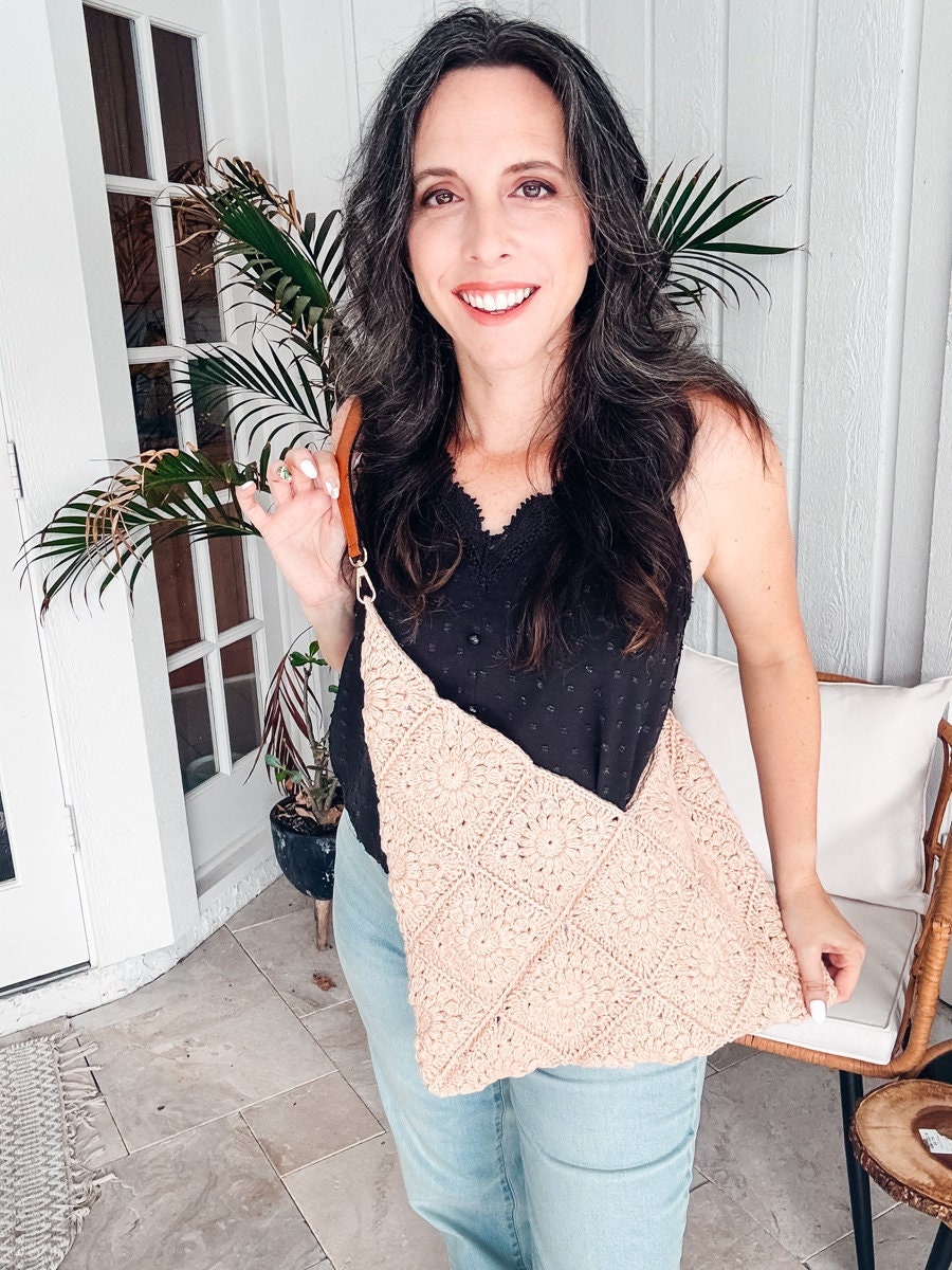 A person with long dark hair smiles while holding a crochet bag, a handmade gift. They are standing indoors near a potted plant, wearing vintage clothes: a black top and light blue pants.