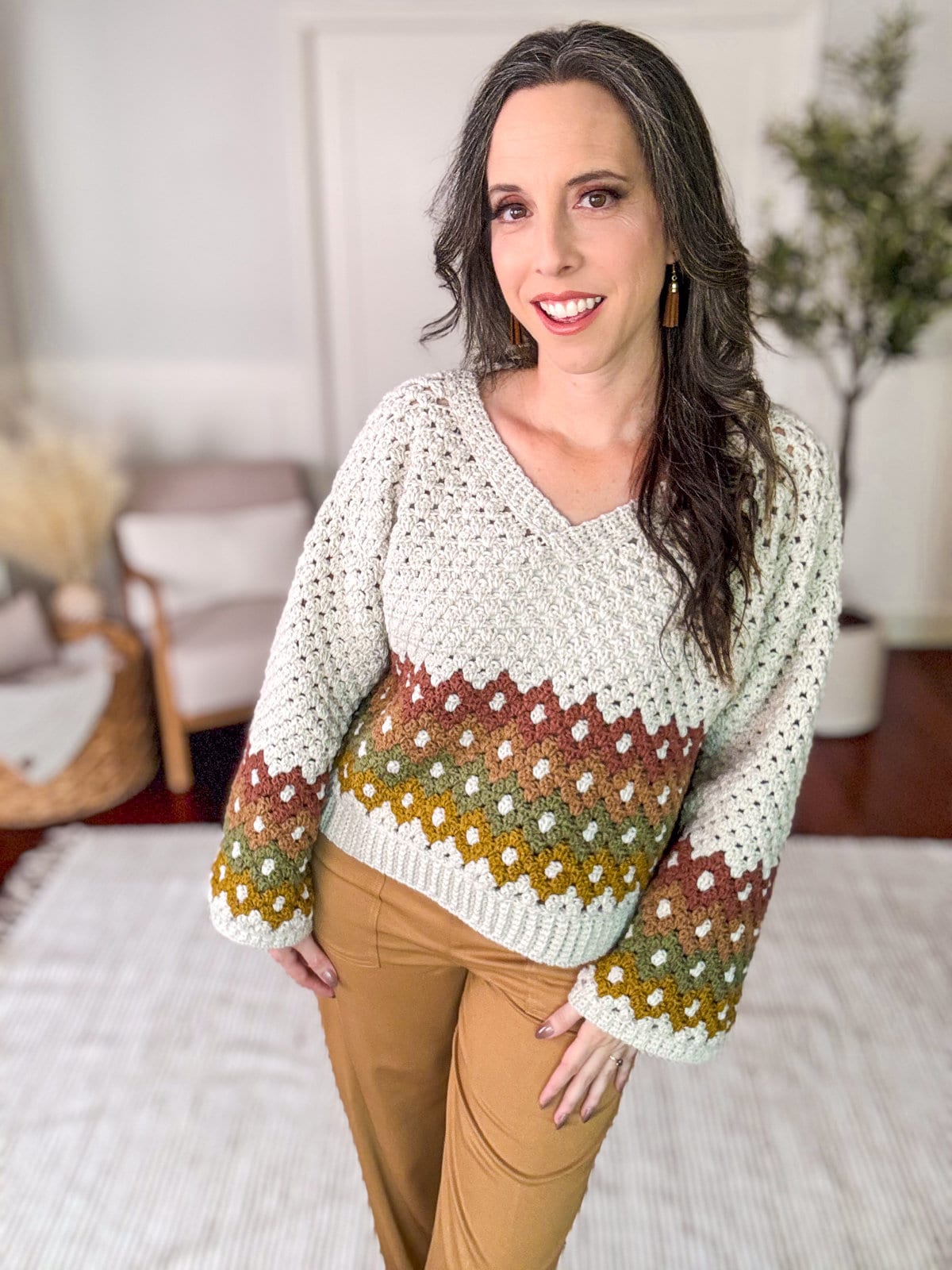 A woman smiles while posing indoors, showcasing her vintage clothes. She is wearing a crocheted sweater with a brown, green, and white pattern, along with tan pants and unique jewellery. There is a plant and furniture in the background.