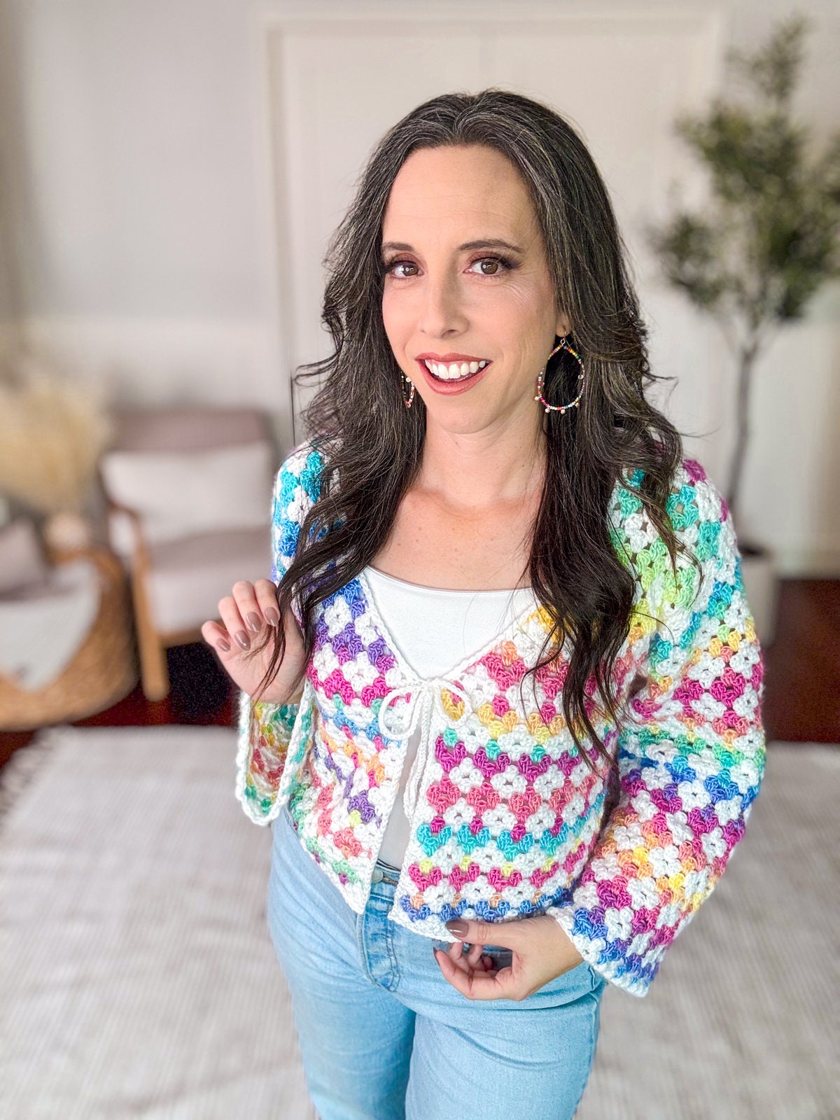 A woman with long dark hair is standing indoors, wearing a colorful crocheted cardigan over a white top and blue jeans. She is smiling and looking at the camera. There are chairs and a plant in the background.