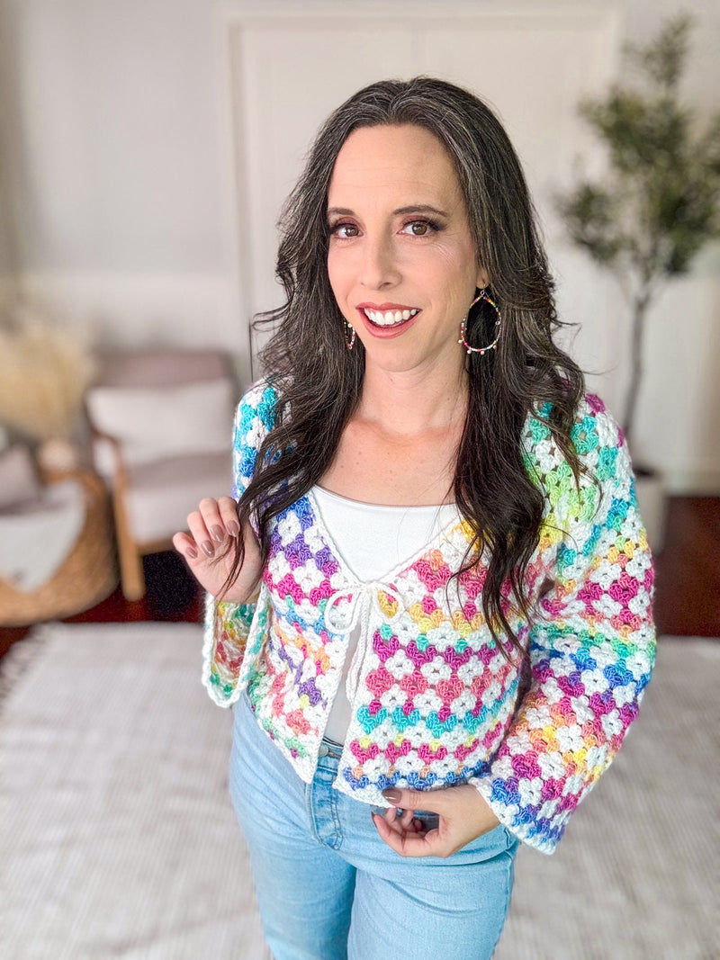 A woman with long dark hair is standing indoors, wearing a colorful crocheted cardigan over a white top and blue jeans. She is smiling and looking at the camera. There are chairs and a plant in the background.