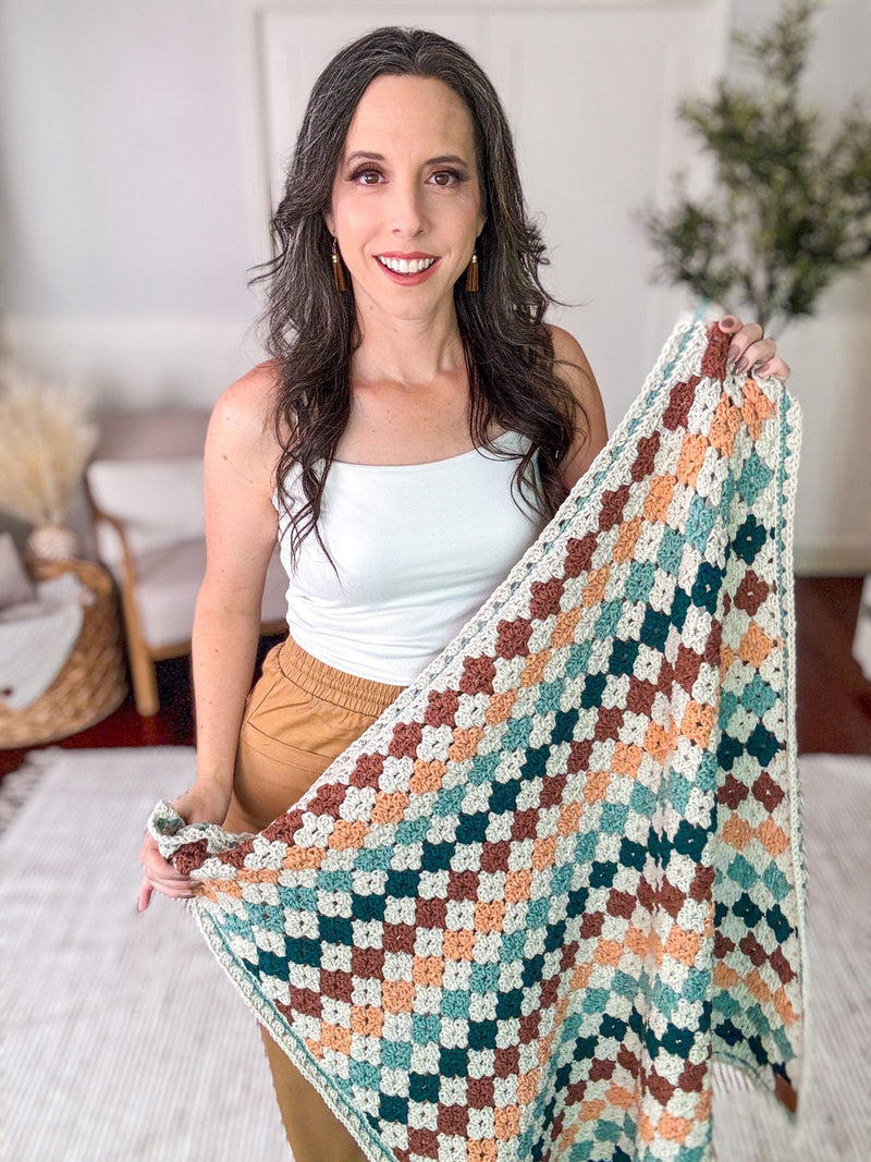 A woman in a white tank top holds a colorful crocheted blanket, standing in a room with a wicker basket and potted plant, surrounded by unique jewellery.