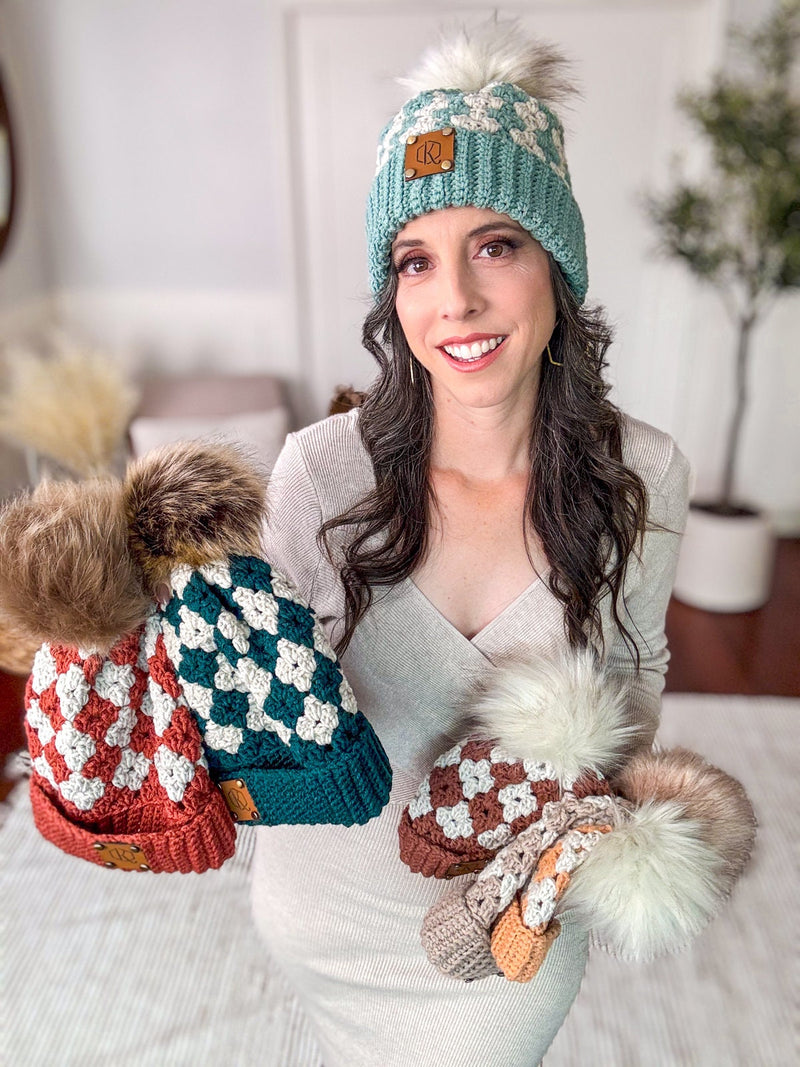 A woman stands indoors holding three crocheted hats in various colors. She is wearing a matching crocheted hat and smiling at the camera, looking as if she’s just received a cherished handmade gift.
