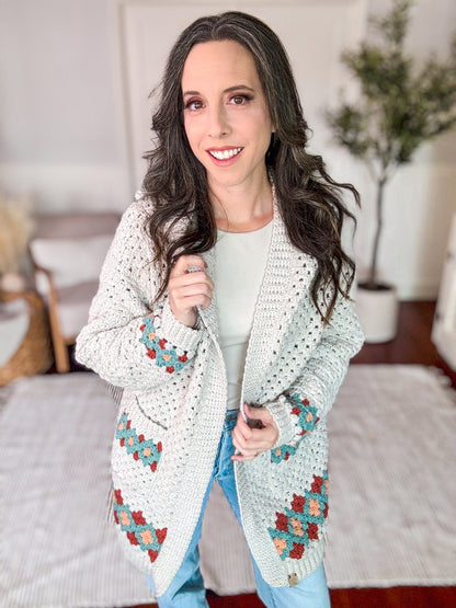 A woman with long brown hair stands indoors, smiling and holding the lapels of a crocheted cardigan with colorful patterns. She is wearing a white top, blue jeans, and unique jewellery that complements her vintage clothes perfectly.