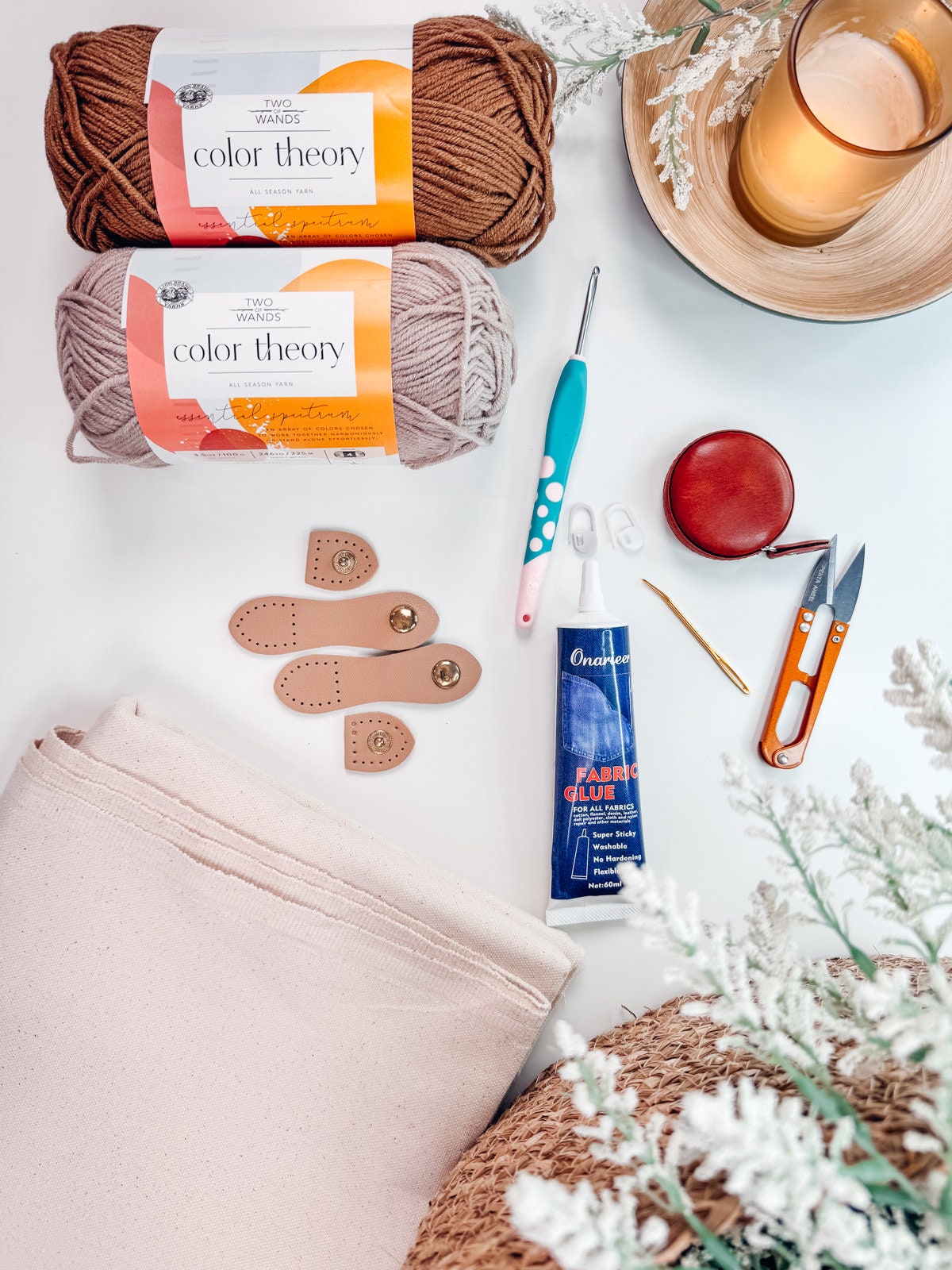 Flat lay of crochet supplies including yarn, a crochet hook, fabric, fabric glue, scissors, a measuring tape, stitch markers, a candle, and some decorative foliage on a white background.