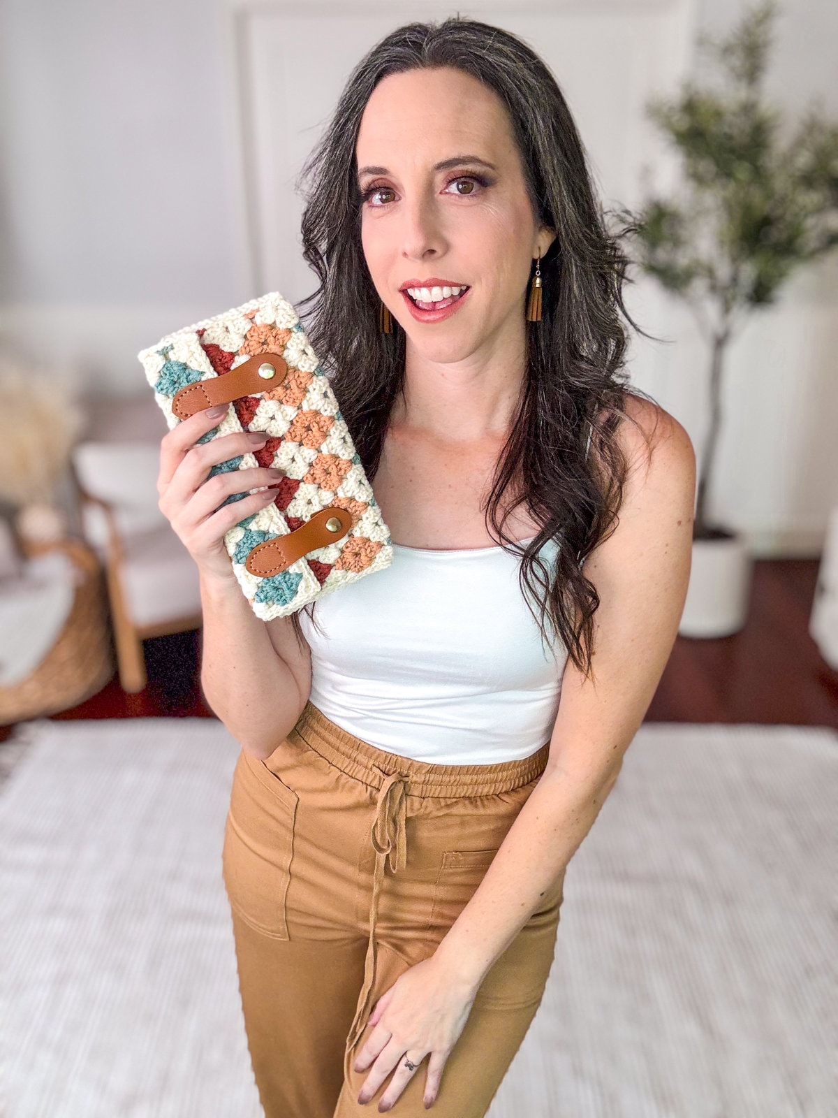 A person holding a crocheted bag with a white tank top and brown pants stands in a living room, smiling at the camera. They exude charm with their vintage clothes and unique jewelry that perfectly complement the handmade gift in their hand.