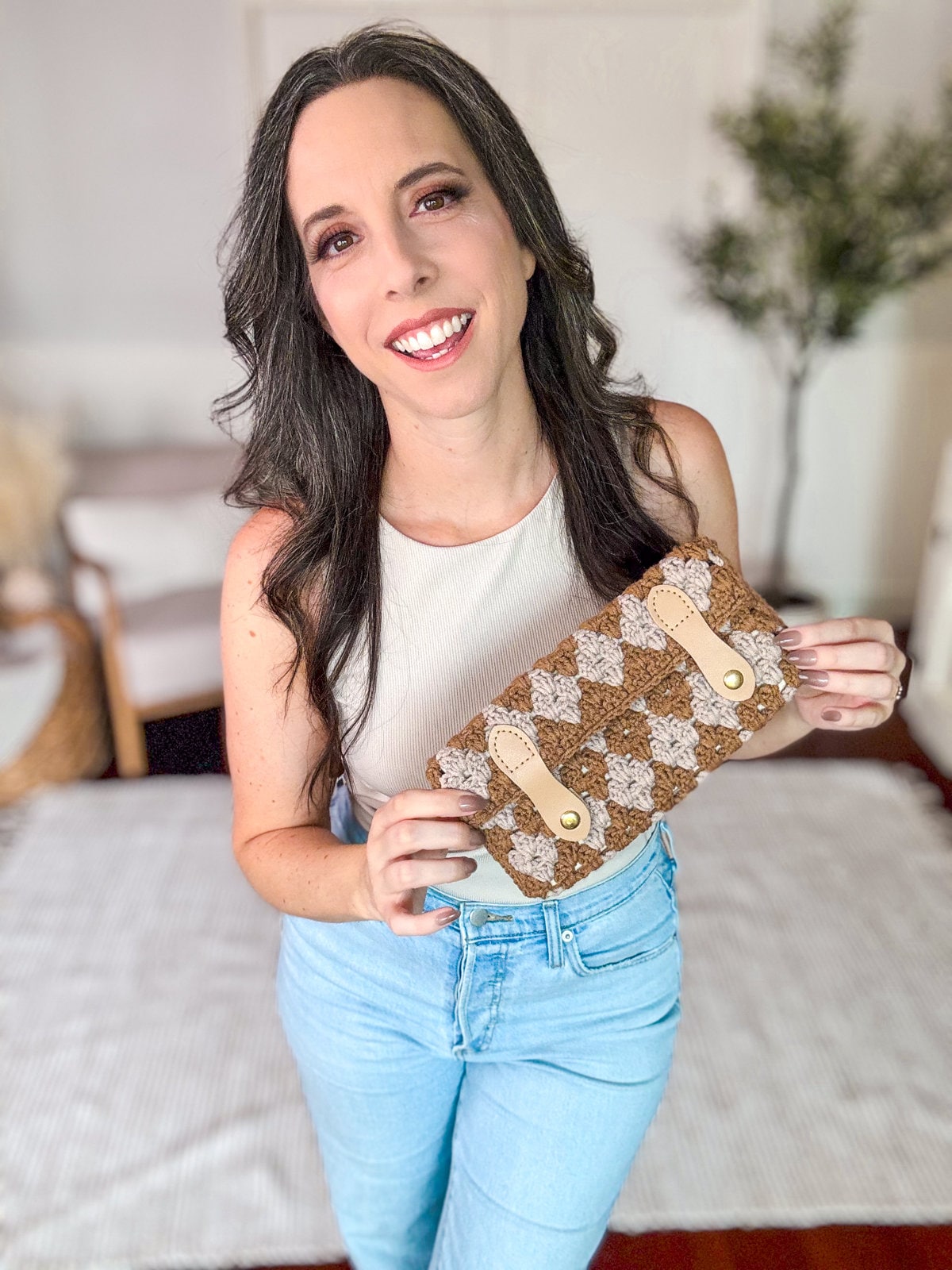 A woman with long dark hair, wearing a sleeveless top and jeans, is smiling and holding a handmade gift—a brown woven clutch bag—while standing indoors.